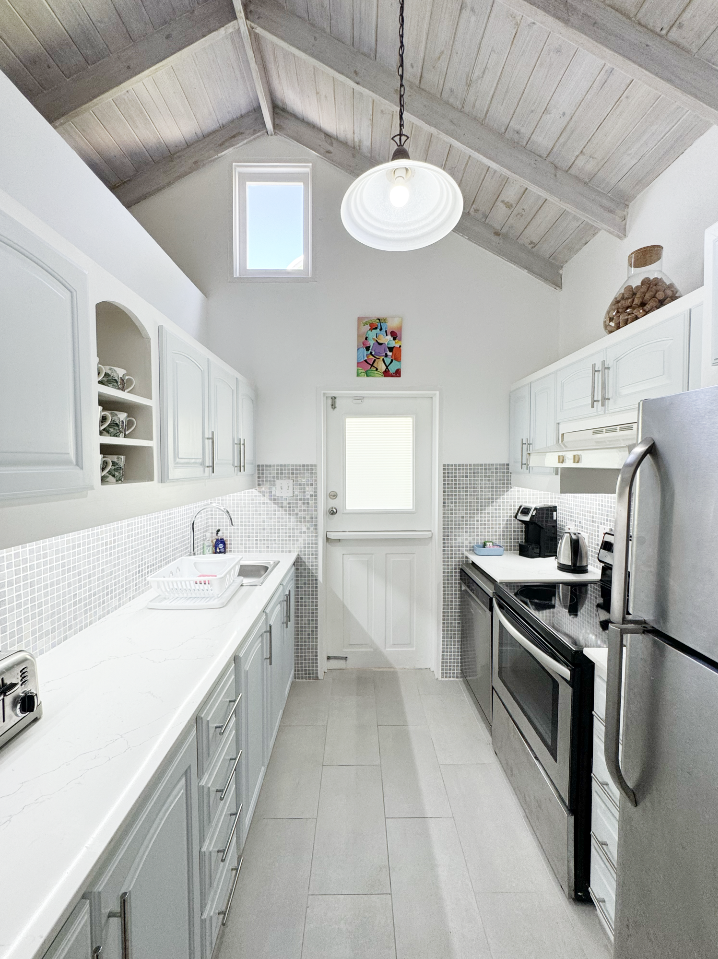 A compact kitchen with white cabinets, a white countertop, small tile backsplash, and stainless steel appliances. The ceiling is vaulted with exposed wooden beams and a skylight, with a pendant light hanging from the ceiling. There are coffee mugs on