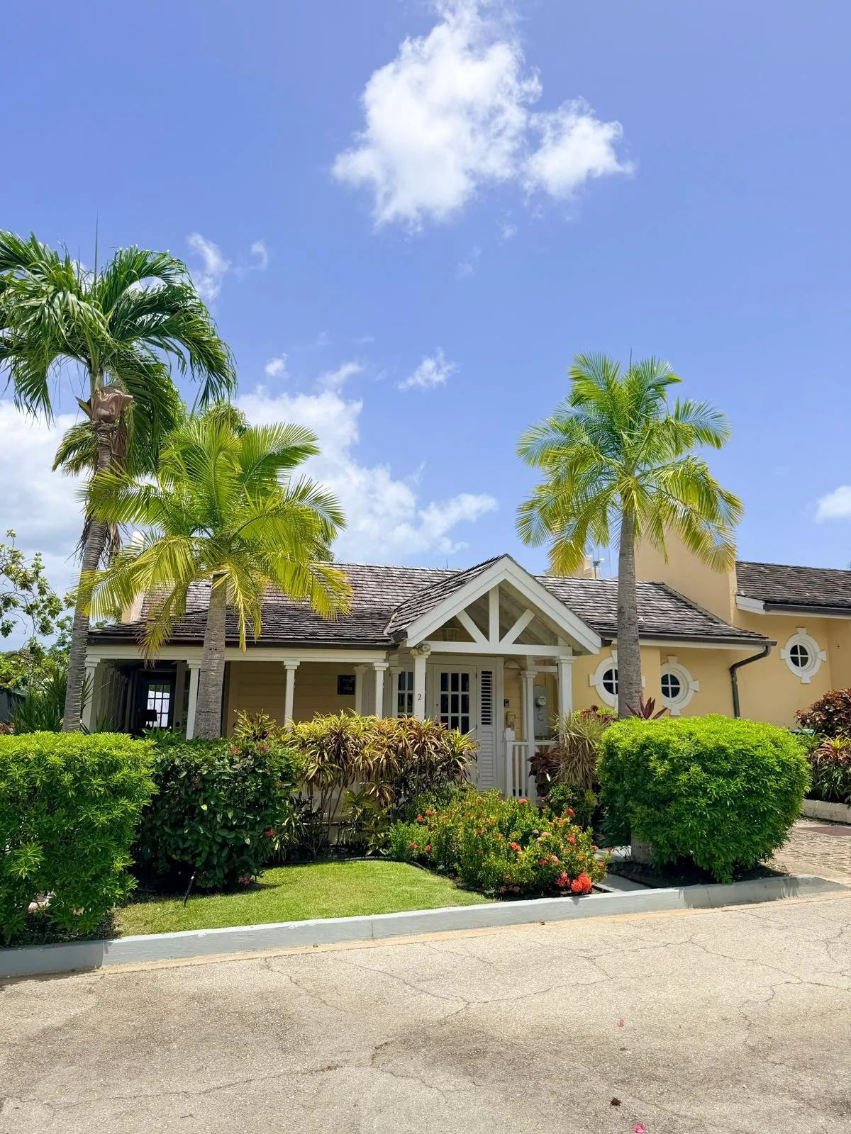 A yellow house with a gabled roof, surrounded by lush green bushes and two tall palm trees, with a bright blue sky and a few white clouds above.