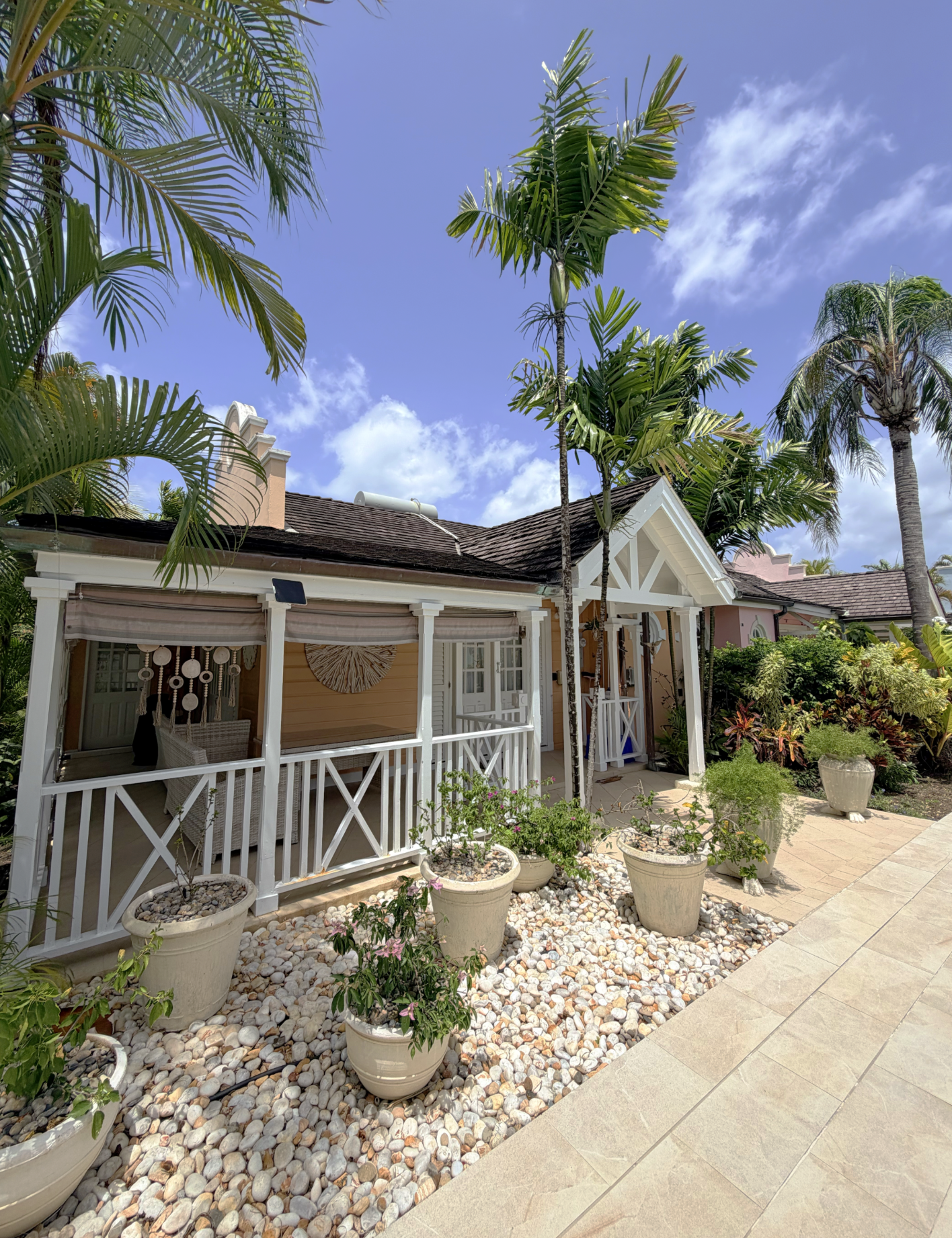 A sunny backyard porch with white railing and potted plants, surrounded by tropical palm trees and lush greenery, under a blue sky with clouds.