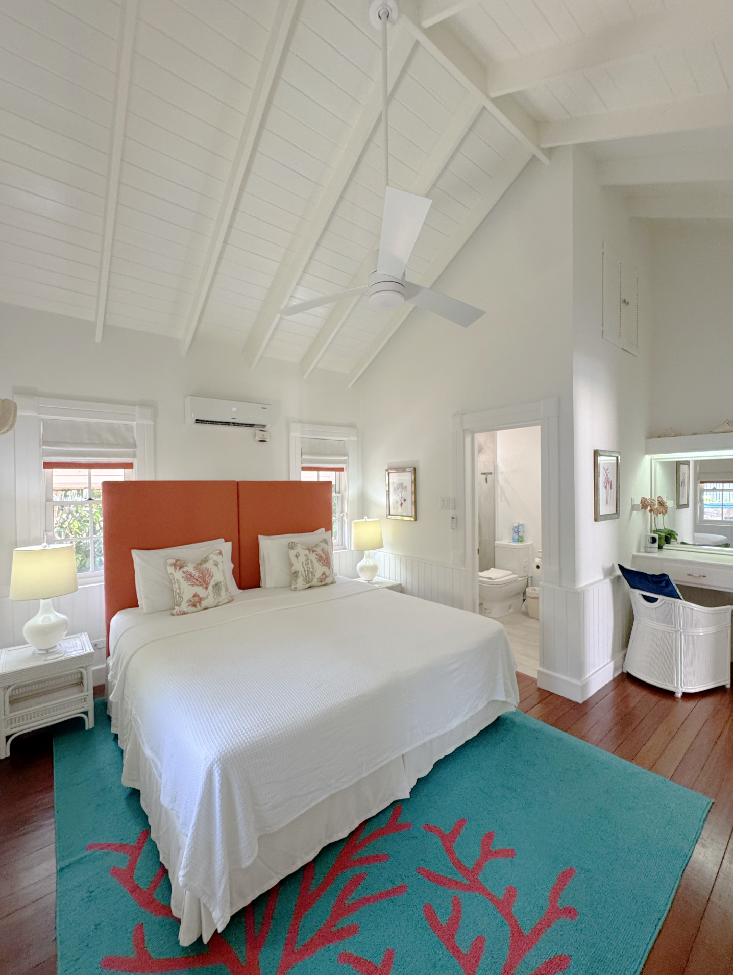Beach-inspired bedroom with white walls, high vaulted ceiling, and wooden floors. Features a large bed with white bedding and coral-themed pillows, an orange headboard, and a blue rug with coral patterns. Two white nightstands with lamps flank the be