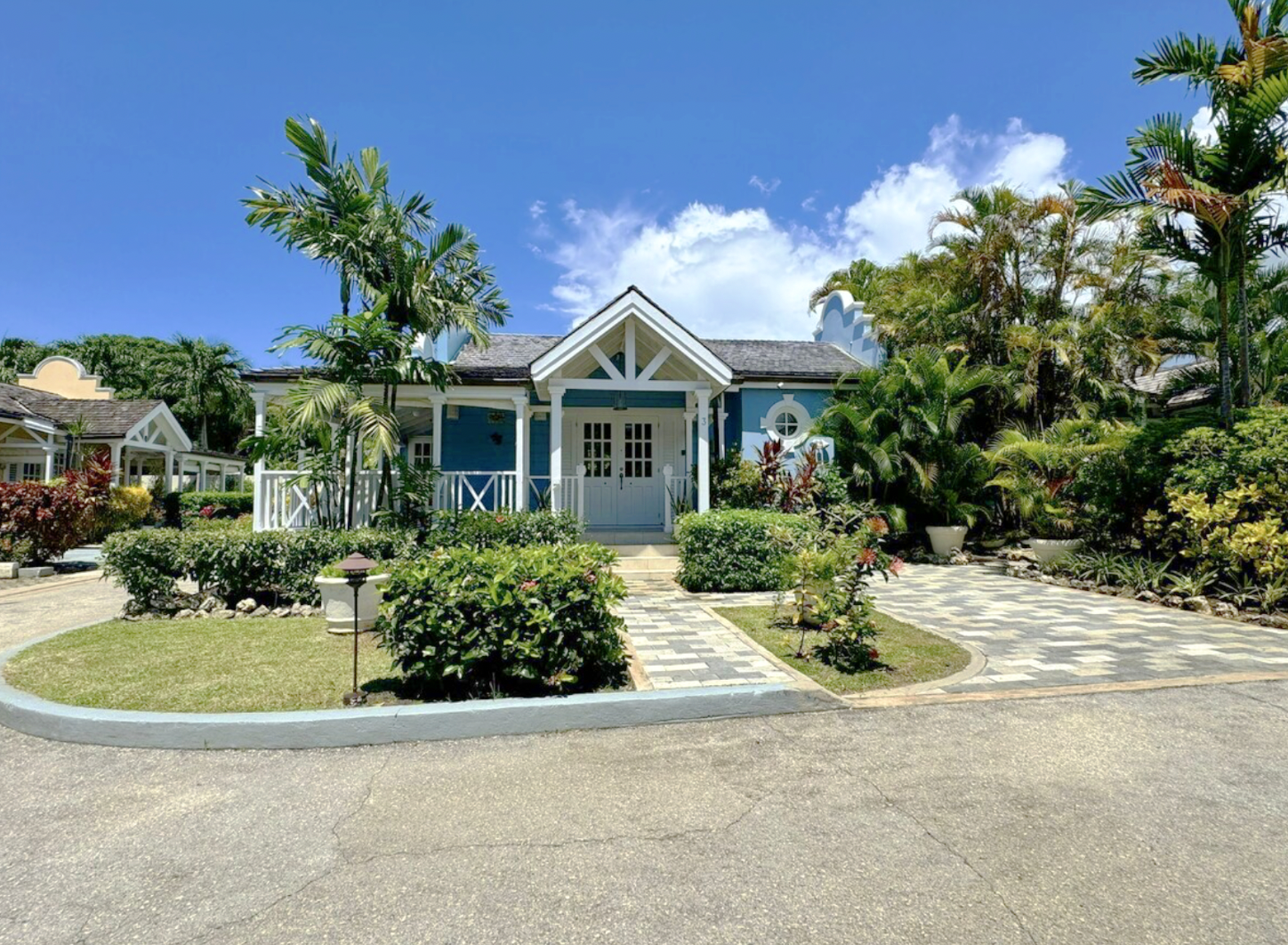 A colorful house with a blue exterior, white trim, and a front porch surrounded by lush tropical plants, palm trees, a guest house, and a paved driveway on a sunny day with a blue sky.