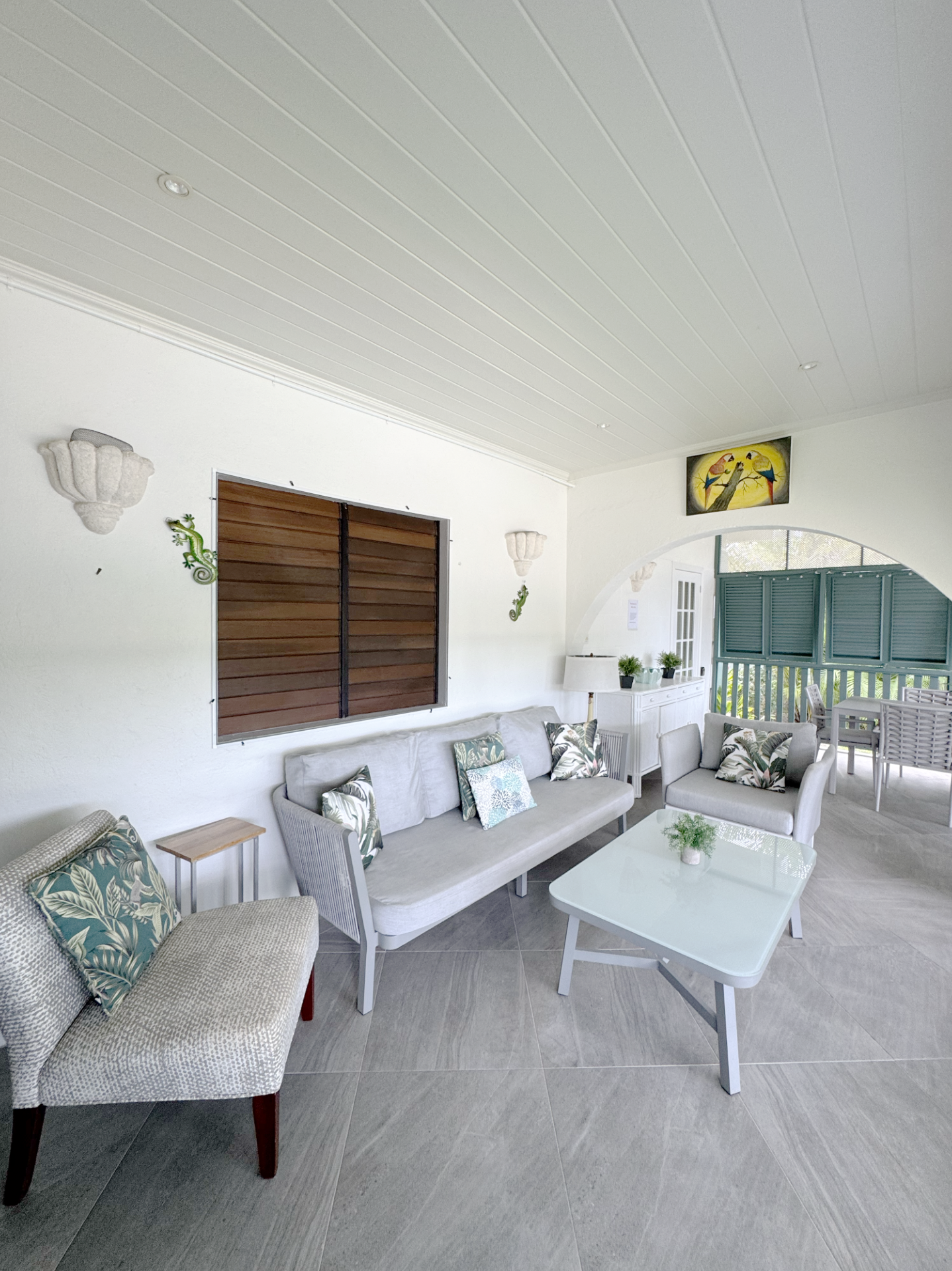 Living room with white walls and gray tiled floor, featuring beige and patterned chairs, a gray sofa with patterned pillows, a white coffee table with a small green plant, a window with wooden shutters, a white sideboard with plants, and a colorful p