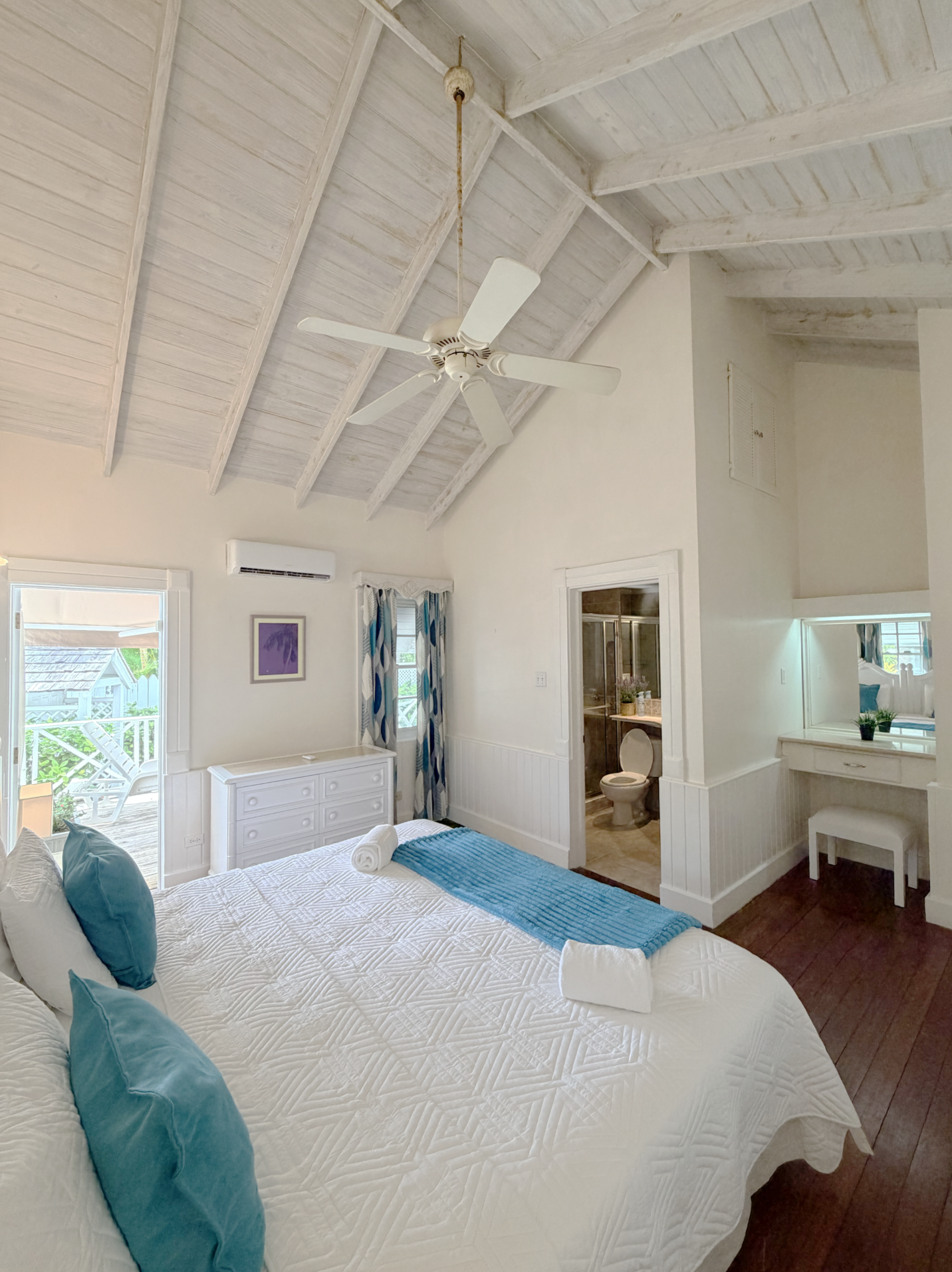 A bedroom with a white bed, teal pillows, and a blue blanket. The room has a vaulted ceiling with a ceiling fan, a window with patterned curtains, and a small writing desk with a mirror. An adjoining bathroom with a toilet or bidet and a shower is vi