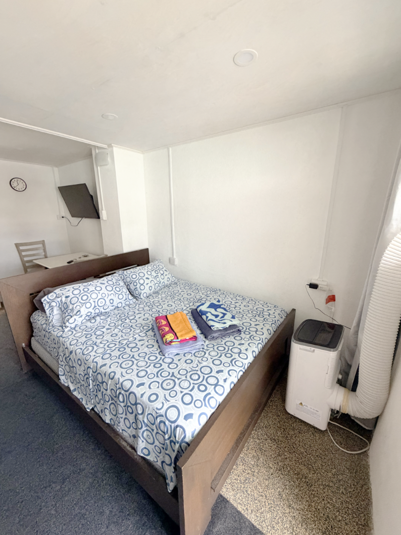 Hotel room with a double bed covered in blue and white circle patterned sheets, with three folded towels on top, a mini air conditioner near the window, a wall-mounted TV, a small table with two chairs, a clock on the wall, and white walls.