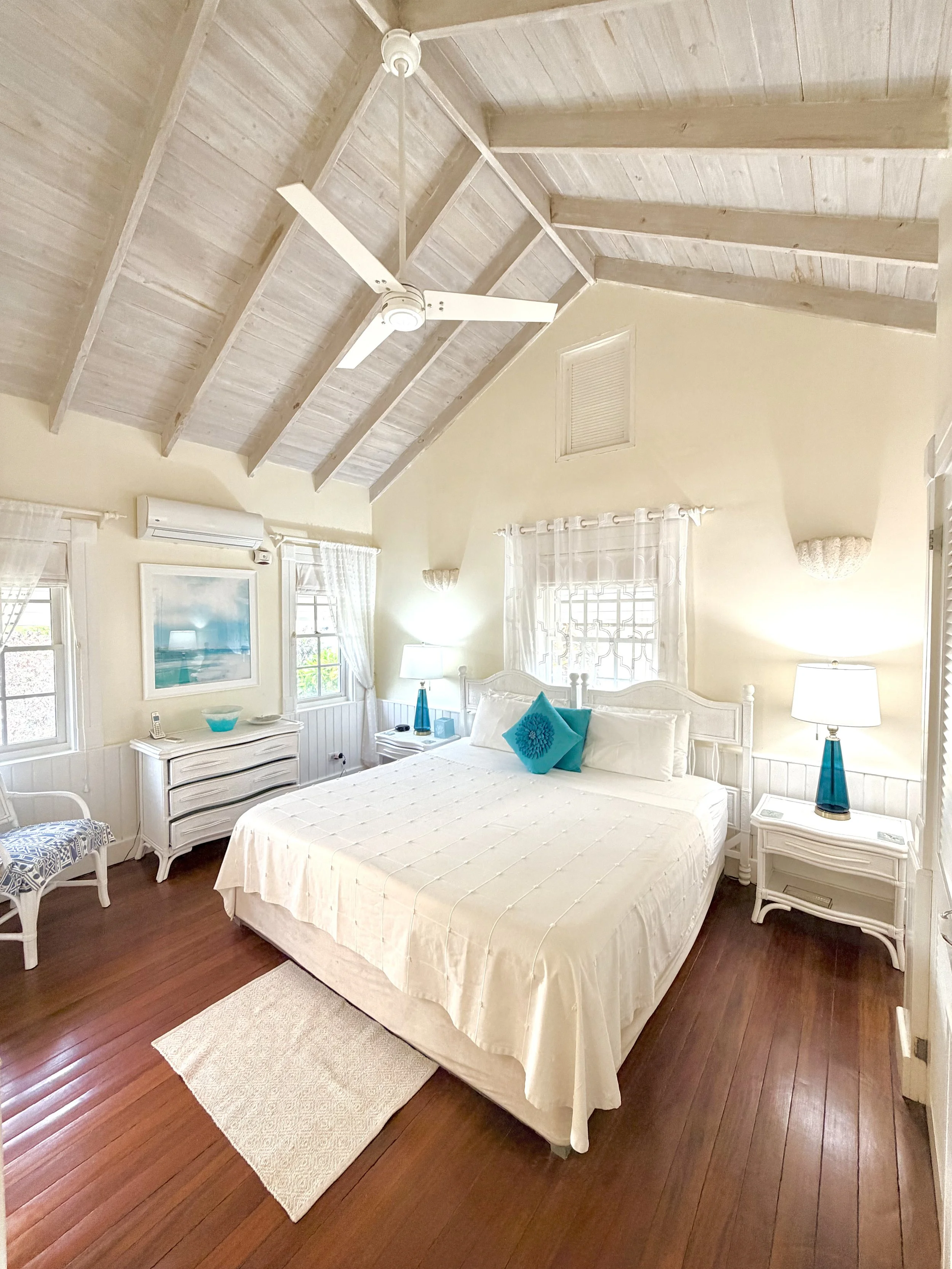 A bedroom with a white bed, turquoise pillows, white furniture, a ceiling fan, and hardwood floors.