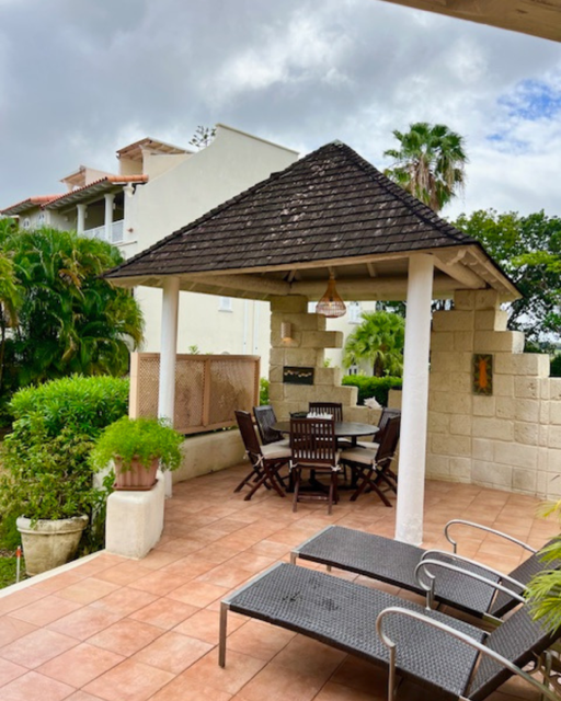 Outdoor patio area with a picnic table under a gazebo and lounge chairs on tiled flooring, surrounded by greenery and residential buildings.