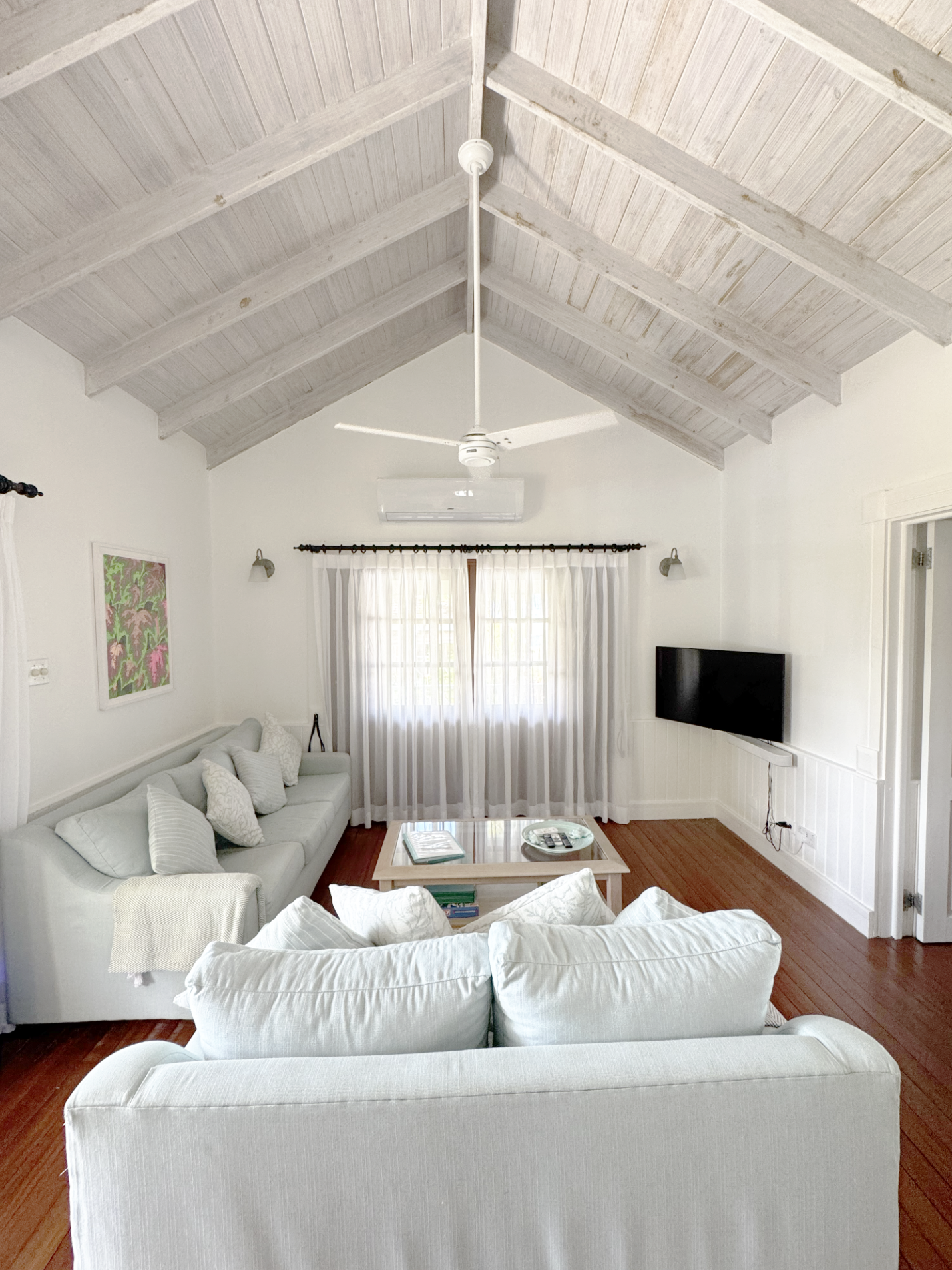 A living room with white sofas, a wooden coffee table, a wall-mounted flat-screen TV, warm wooden flooring, white curtains, and a wooden ceiling with exposed beams and a ceiling fan.