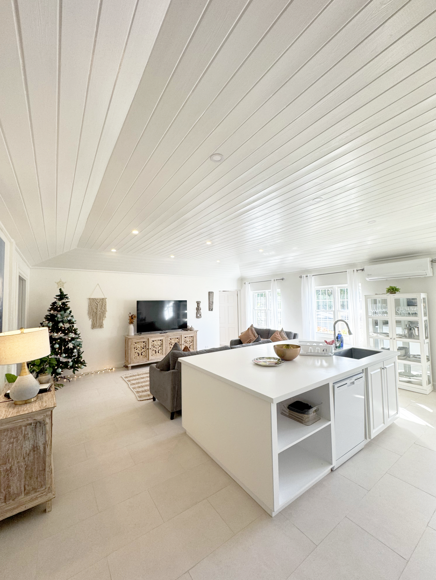 Brightliving room with a white ceiling featuring recessed lighting, a decorated Christmas tree, a large flat-screen TV on an ornate console, a grey sofa with pillows, a wooden side table with a lamp, a white kitchen island with a bowl and dishes, and