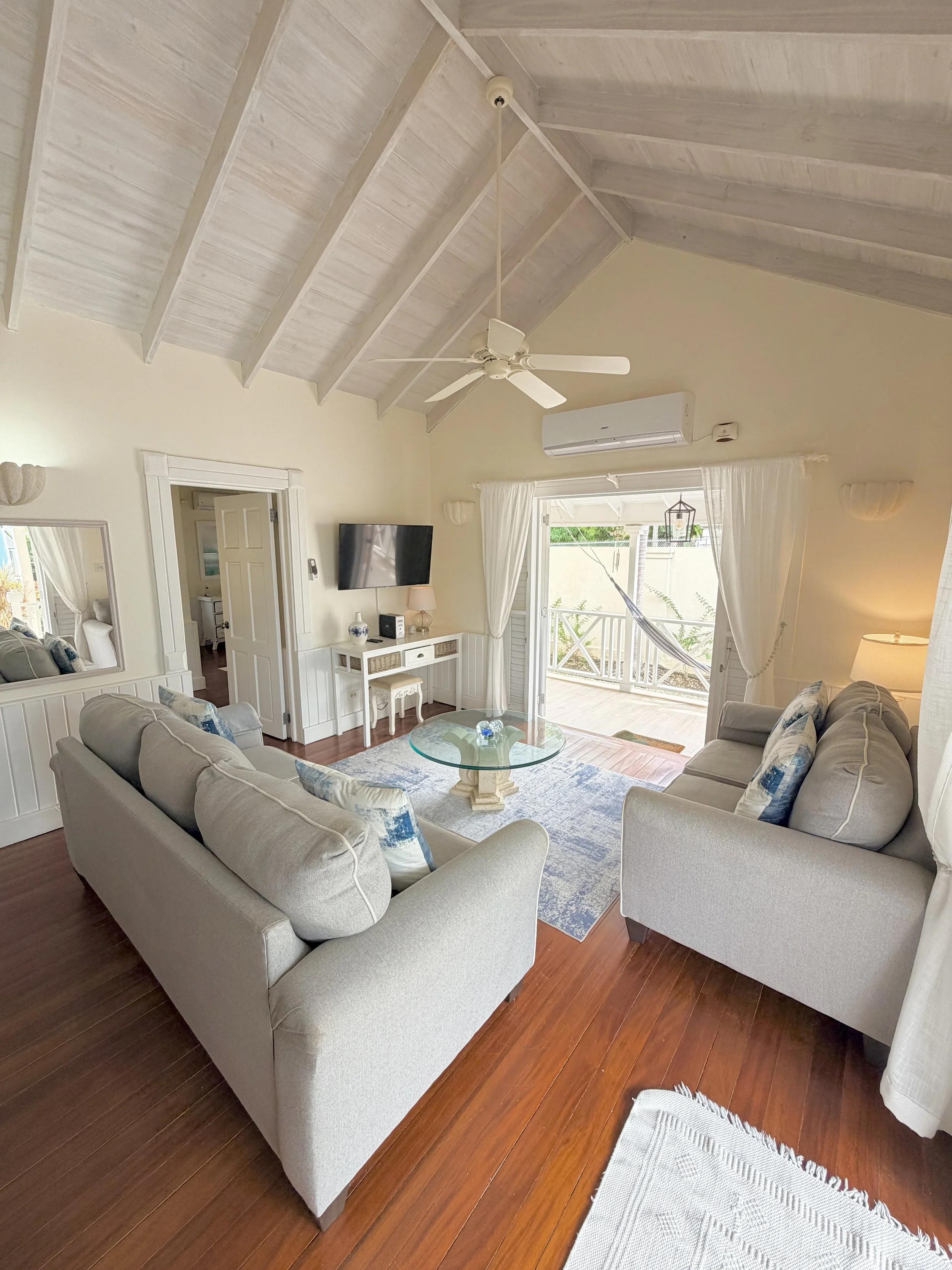 A living room with two beige sofas arranged in a U-shape around a glass coffee table, a white TV mounted on the wall, a doorway leading to another room, and a balcony with a glass door and curtains letting in natural light.