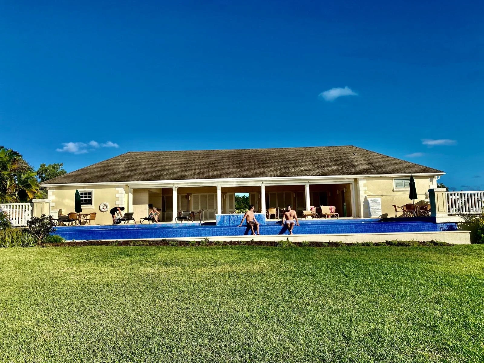 People swimming and lounging around a blue swimming pool in front of a single-story building with a brown shingle roof, outdoor furniture, and umbrellas.