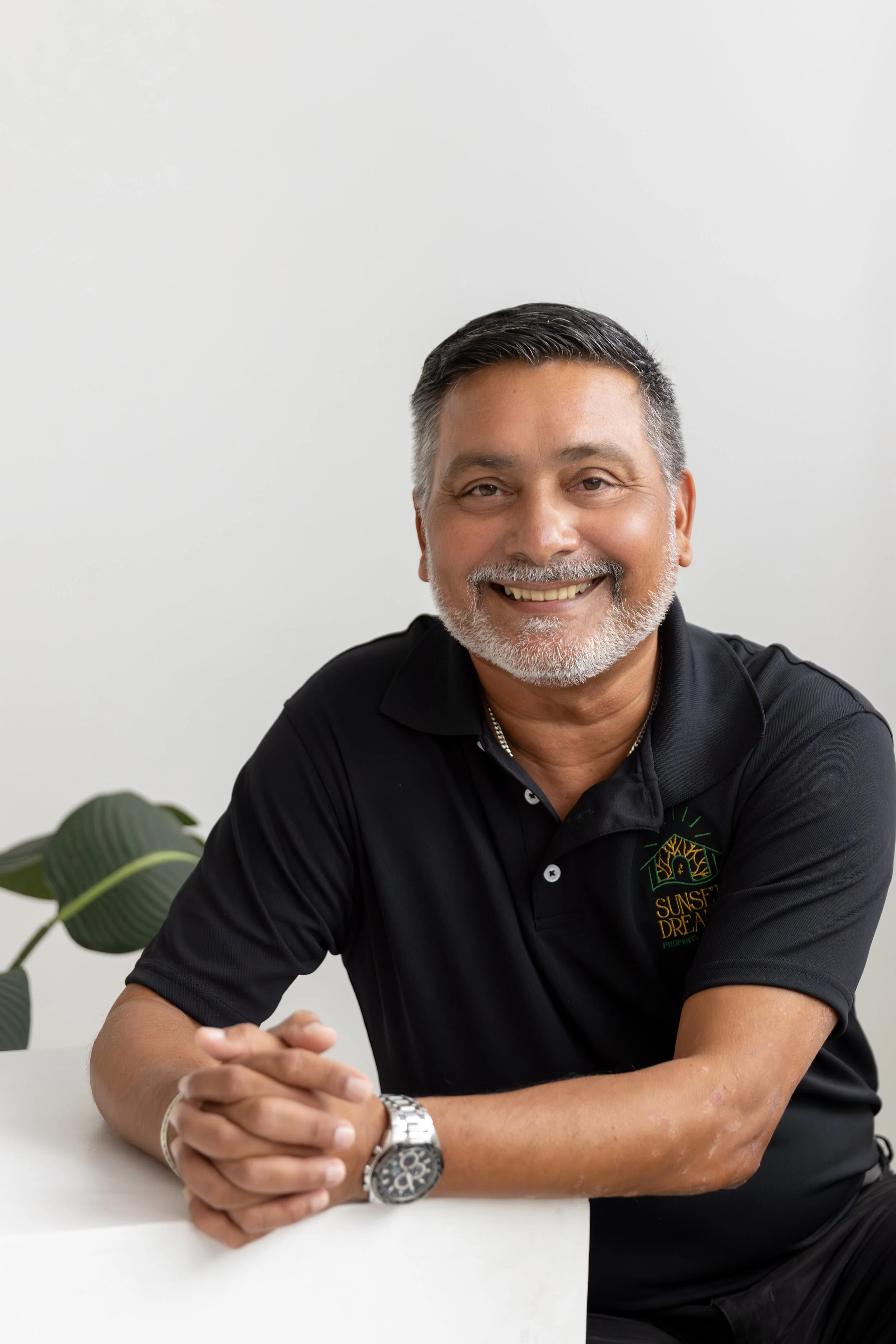 A middle-aged man with short, dark and gray hair, and a beard and mustache, smiling and sitting at a table with his hands clasped. He is wearing a black polo shirt with a logo that reads 'Sunset Dream Property.' There is a green leafy plant visible in the background against a plain white wall.