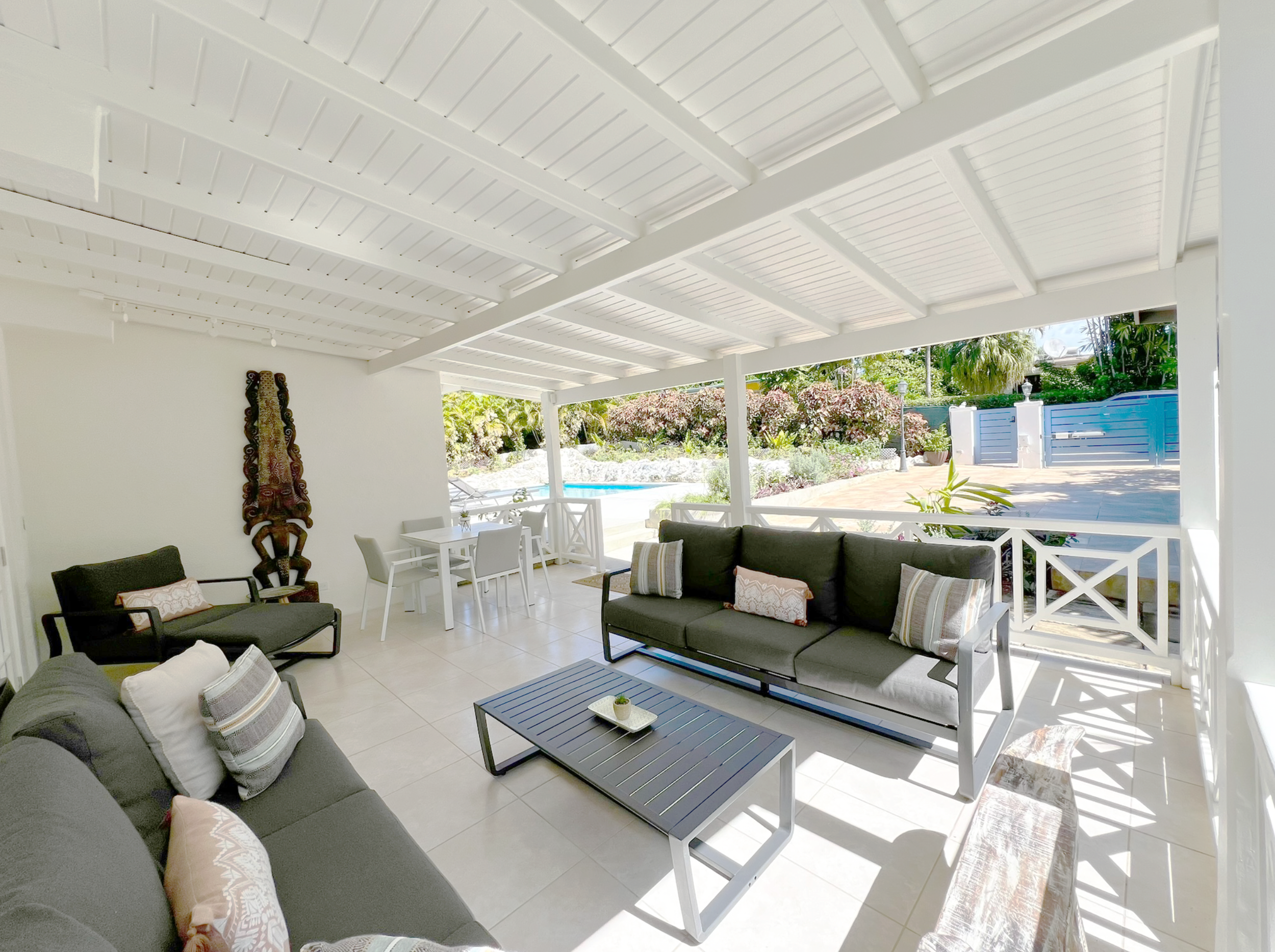 Covered patio area with white wooden ceiling, dark outdoor sofas with pillows, white dining table and chairs, and views of a garden with bushes, palm trees, and a swimming pool.