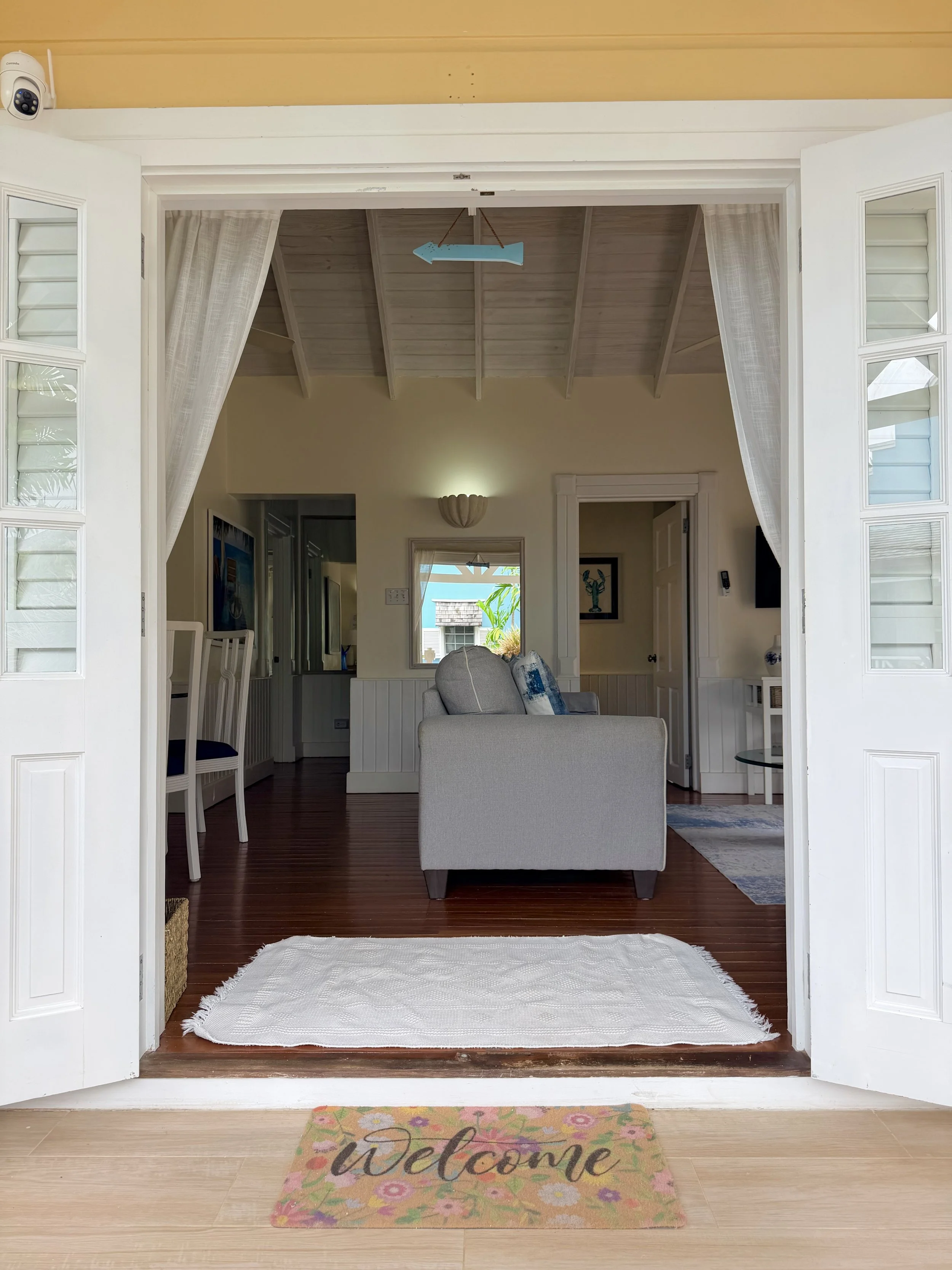 View of an open door leading into a cozy living space with a white couch, wooden flooring, a colorful welcome mat, and several windows allowing natural light, decorated in a beach house style.