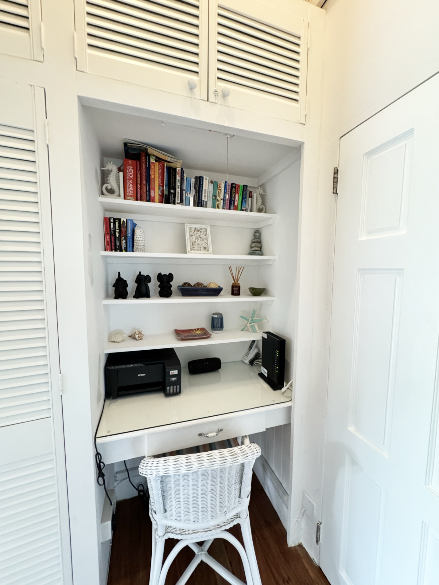 A small home office nook with a white desk, white wicker chair, and shelves filled with books and decorations, located between closet doors and near a wall with a door.