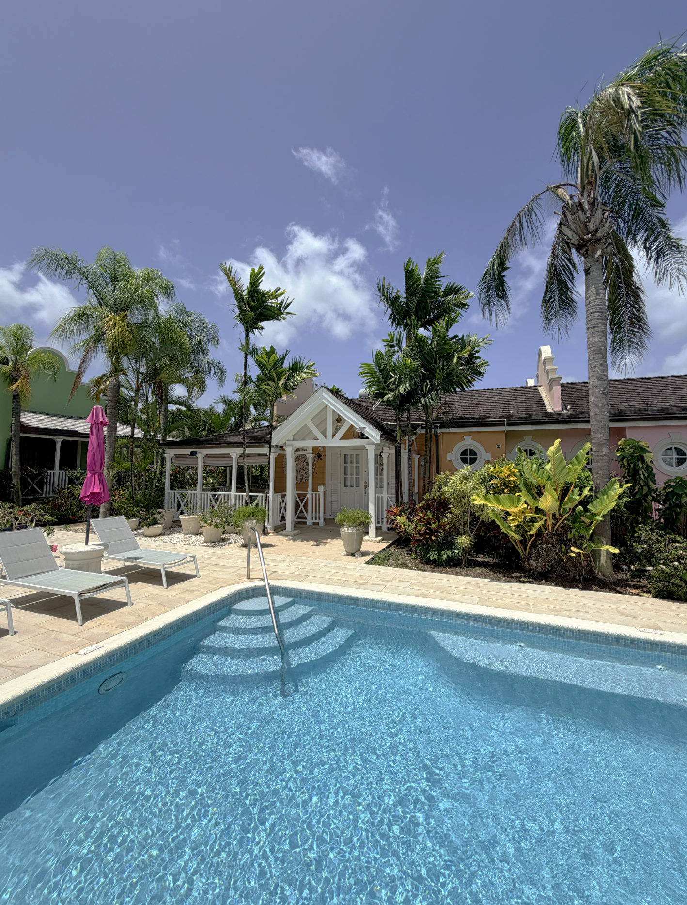 A backyard with a swimming pool, surrounded by lounge chairs and a pink umbrella, with palm trees and a yellow house in the background under a blue sky.