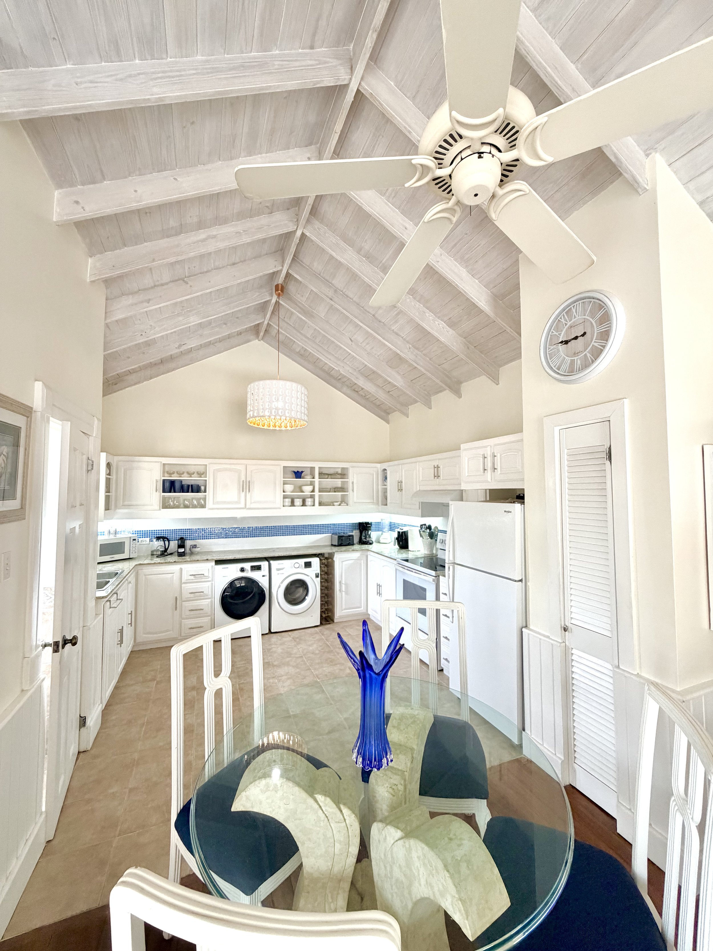 A kitchen with white cabinets, a microwave, washing machines, and a glass dining table with white chairs. A blue glass vase is on the table, with a ceiling fan and clock visible.