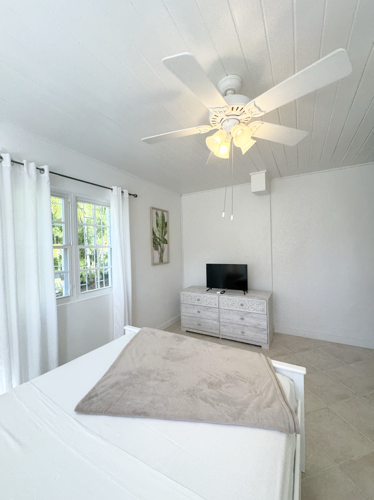 A bedroom with white walls and curtains, a window with greenery outside, a white dresser with a TV on top, and a ceiling fan with lights.