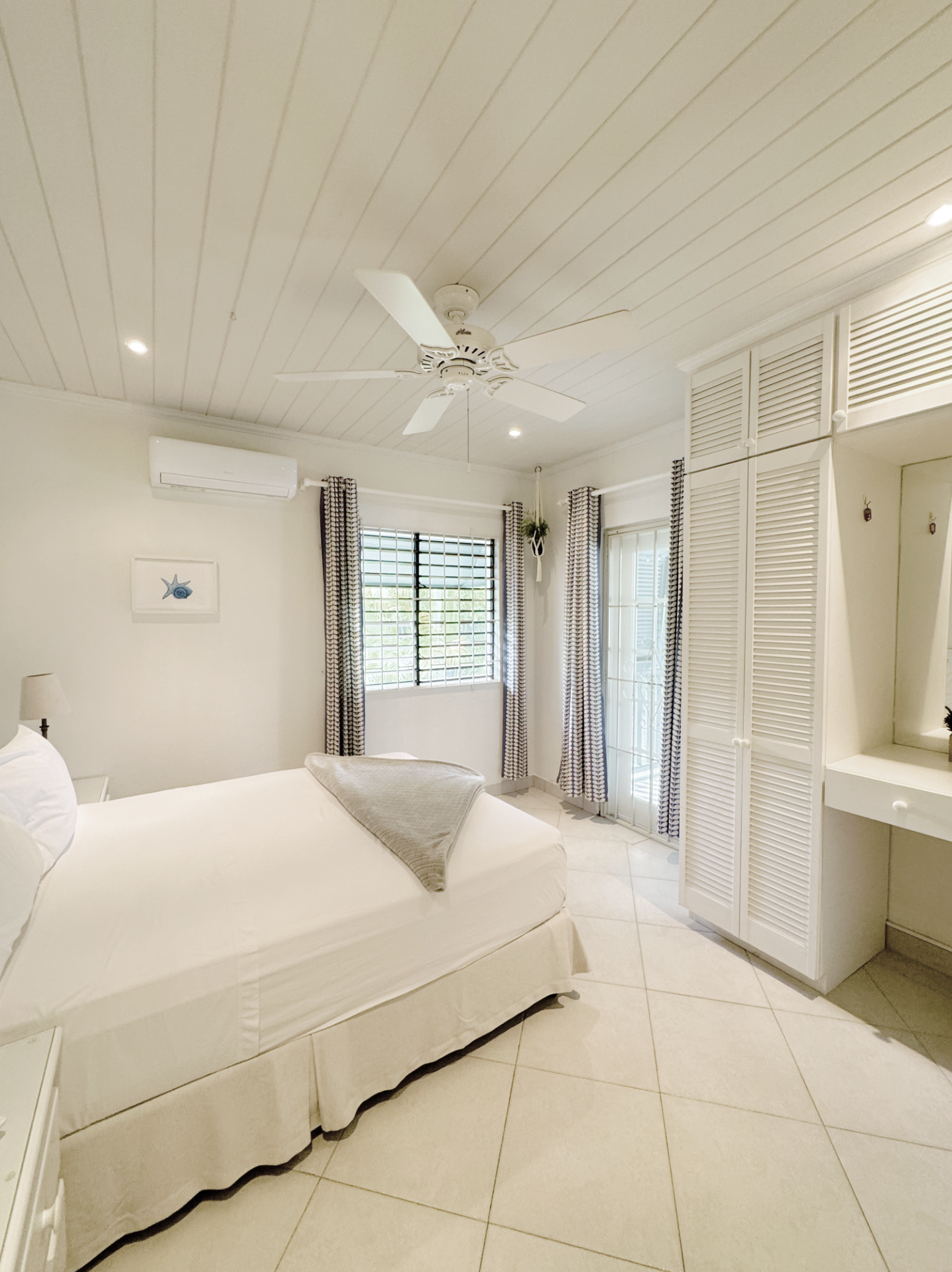 A bedroom featuring a white bed with a sloped skirt, light-colored tiled floor, white paneled ceiling with a ceiling fan, and walls with windows and white louvered closet doors.