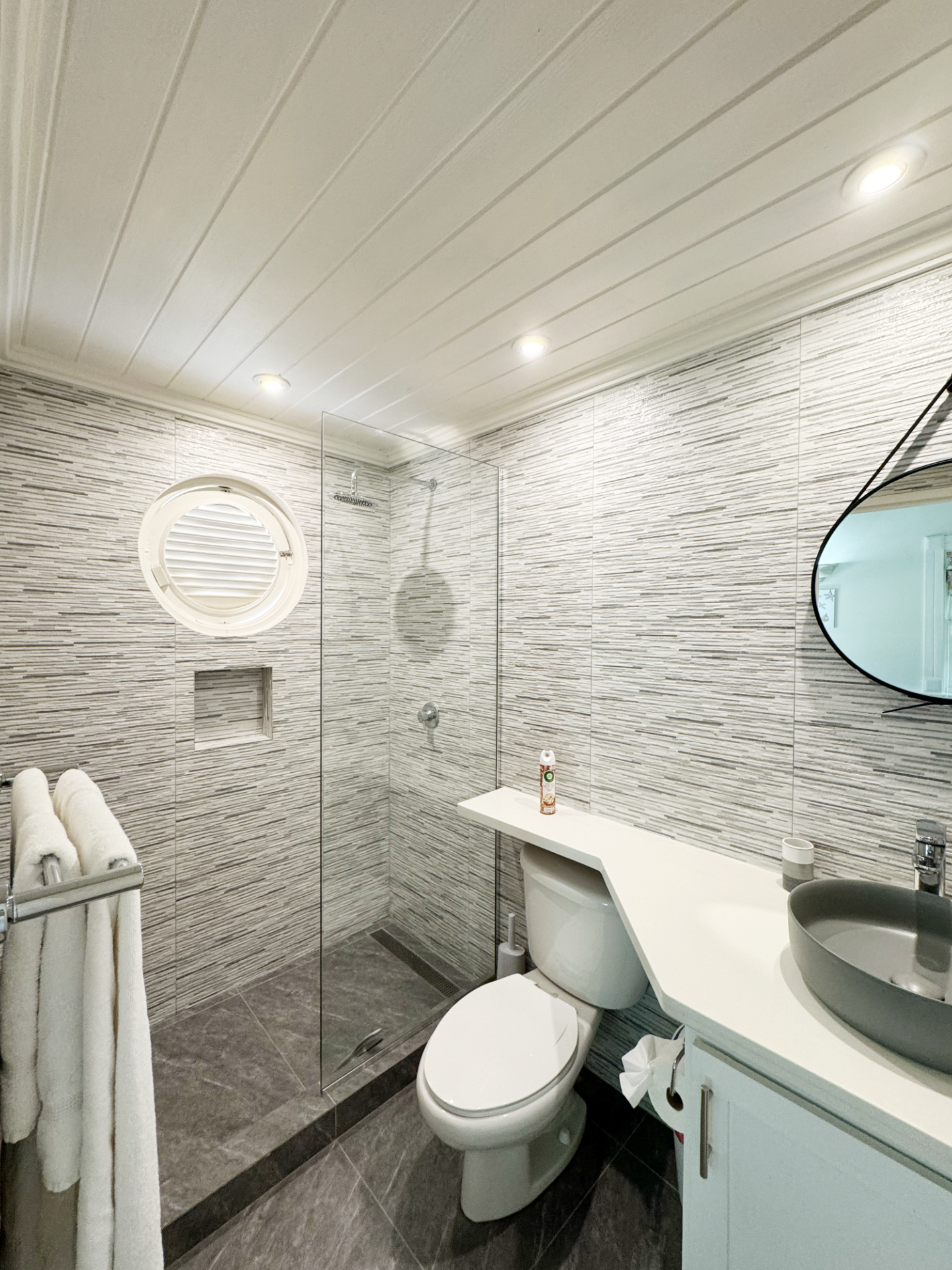 Modern bathroom with a glass shower enclosure, mounted showerhead, and textured wall tiles. Includes a toilet, wall-mounted cabinetry with a countertop, round mirror, and a towel rack with white towels. Recessed lighting on the ceiling.