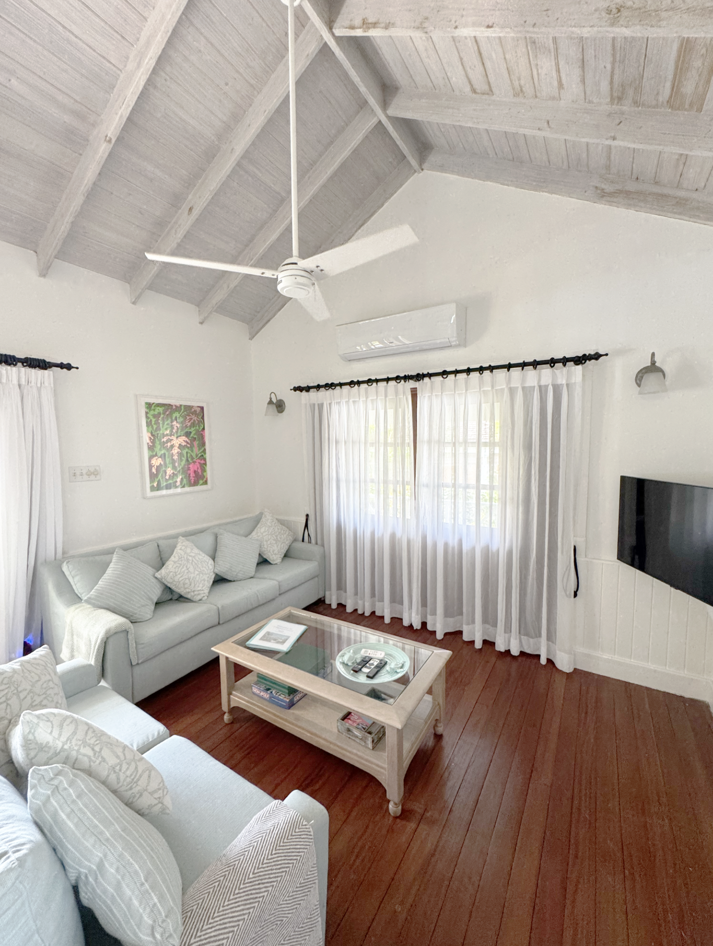 Living room with white ceiling, wooden floor, white sofa with pillows, wooden coffee table with remote controls, books, and an ashtray, white curtains on windows, wall-mounted air conditioner, ceiling fan, framed artwork, and wall sconces.