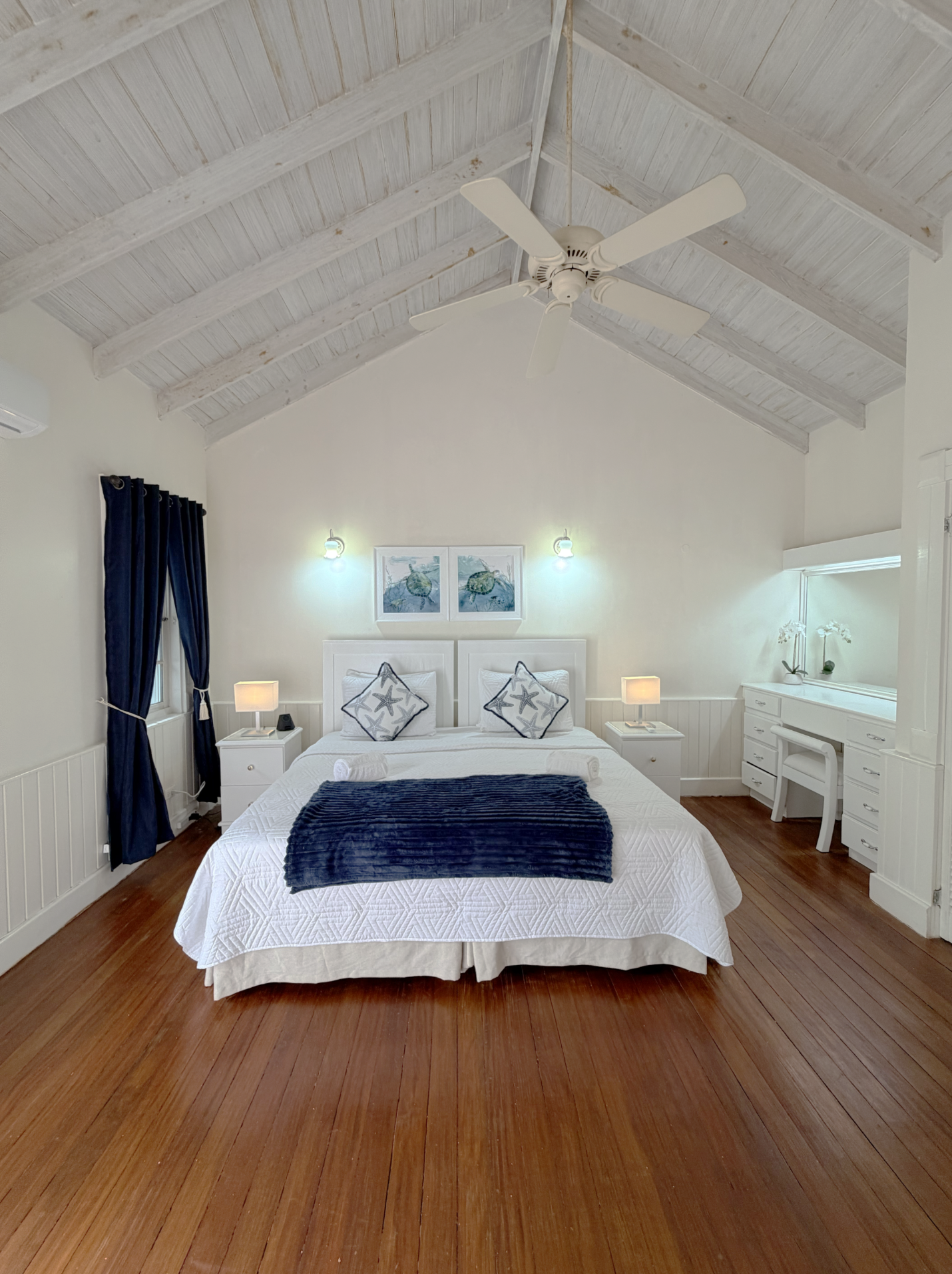 A bedroom with a white bed, navy blue blanket, and starfish pillows, two nightstands with lamps, framed ocean-themed art, dark blue curtains, white dresser, and a ceiling fan with white blades, ceiling made of light-colored wooden planks.