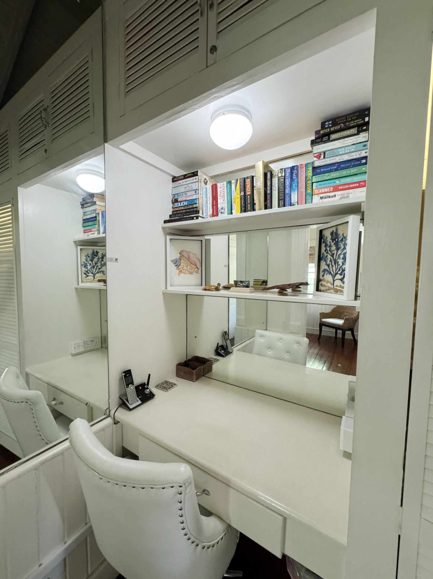 A white office or reception desk with two white cushioned chairs, a mirror, a small white table, and a wall with books and framed artwork. There are two ceiling lights and a large mirror reflecting part of the room.