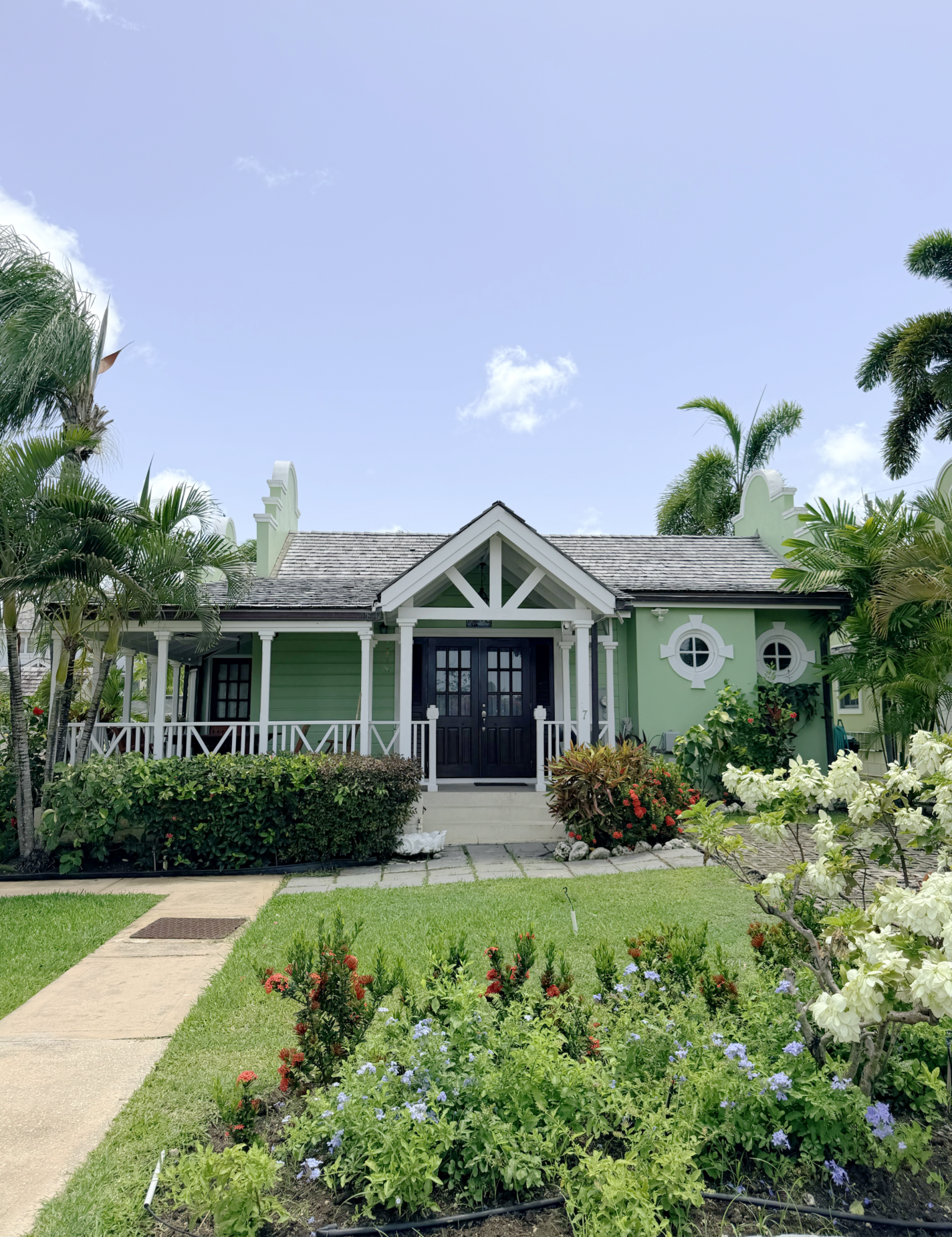Front view of a light green house with black door, surrounded by a well-maintained garden with various flowers and plants, palm trees, and a concrete pathway.