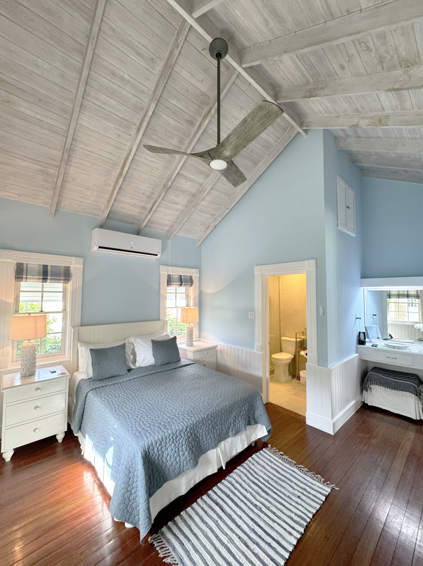 A cozy bedroom with light blue walls, a white headboard, and a gray quilted bedspread. Two windows with striped blinds, a white nightstand with lamps, a small bathroom with a toilet and open door, hardwood floors, a striped rug, and a ceiling fan wit