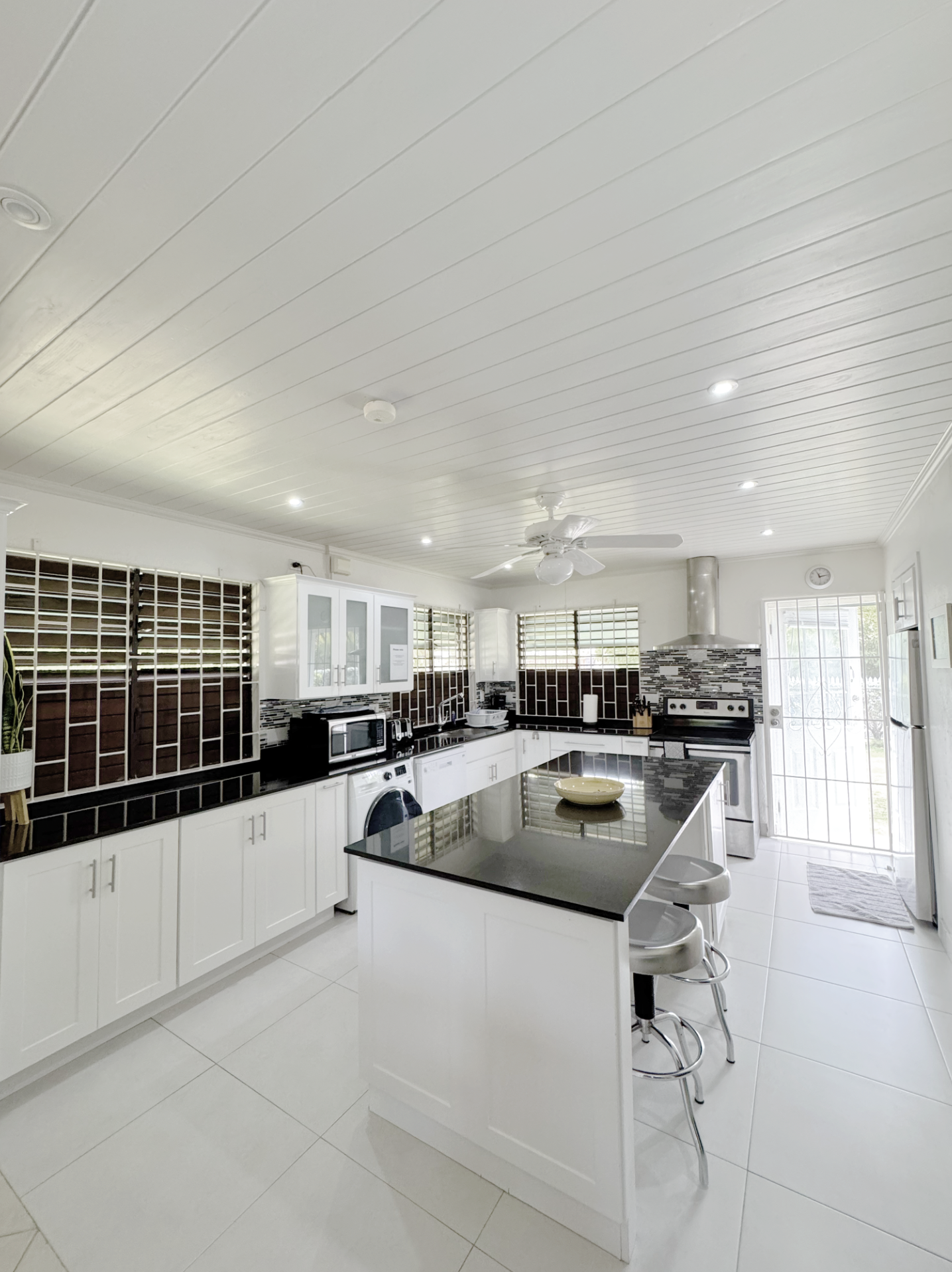 Modern kitchen with white cabinets, black countertops, and a large island with a bowl. Stainless steel appliances including a stove and microwave. Windows with blinds, a clock on the wall, and a ceiling fan. Natural light brightens the room.