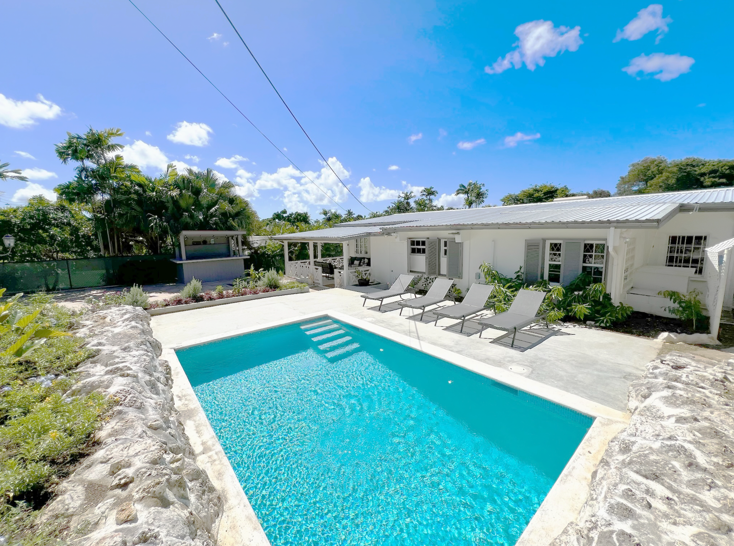 A backyard pool area with a white house, garden chairs, lush green trees, and a bright blue sky with scattered clouds.