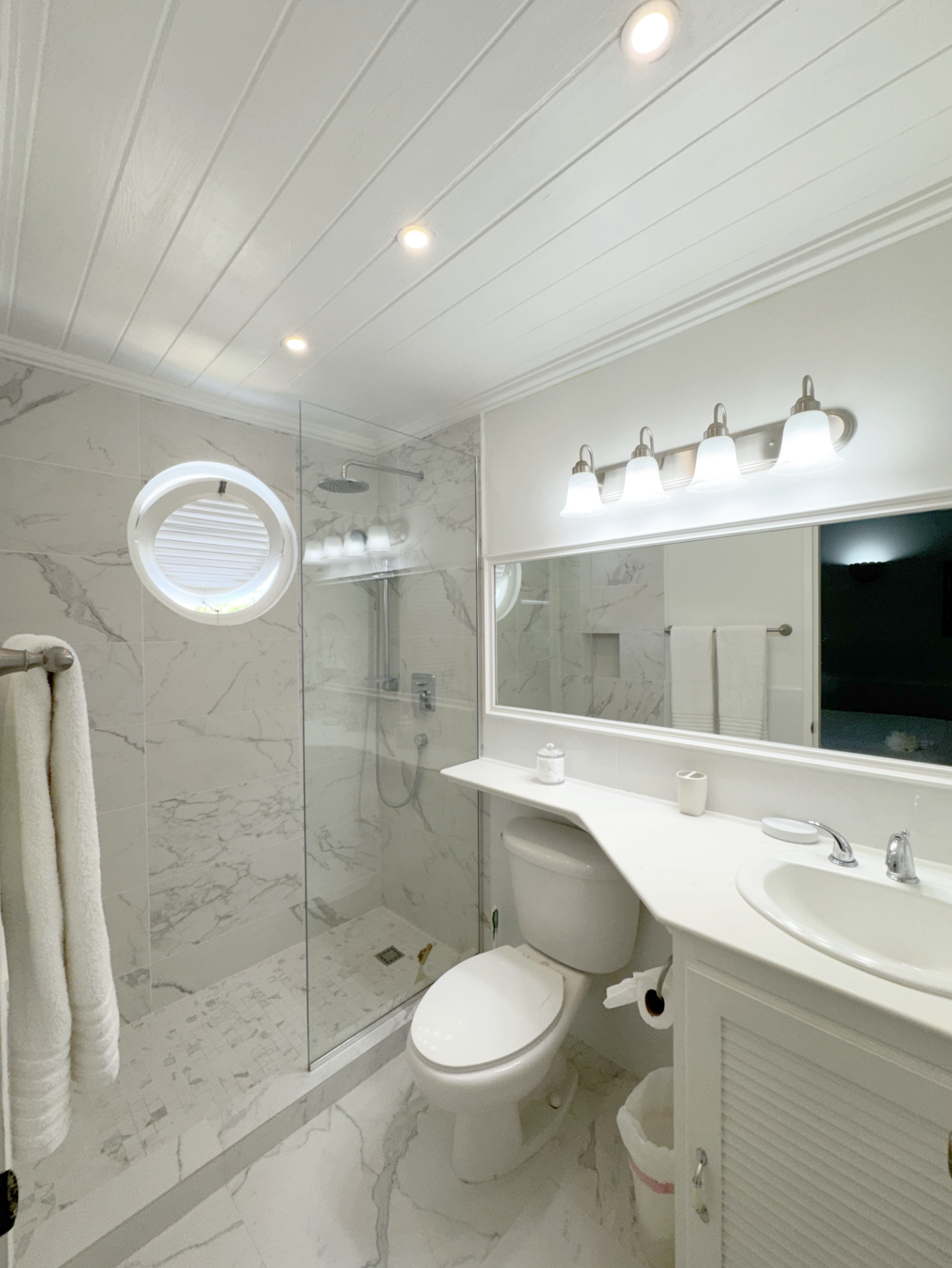 A modern bathroom with a marble-tiled shower with glass doors, a white toilet, a white vanity with a sink, and a large mirror. The bathroom has white beadboard ceiling with recessed lighting and a wall-mounted light fixture with four shades. There is