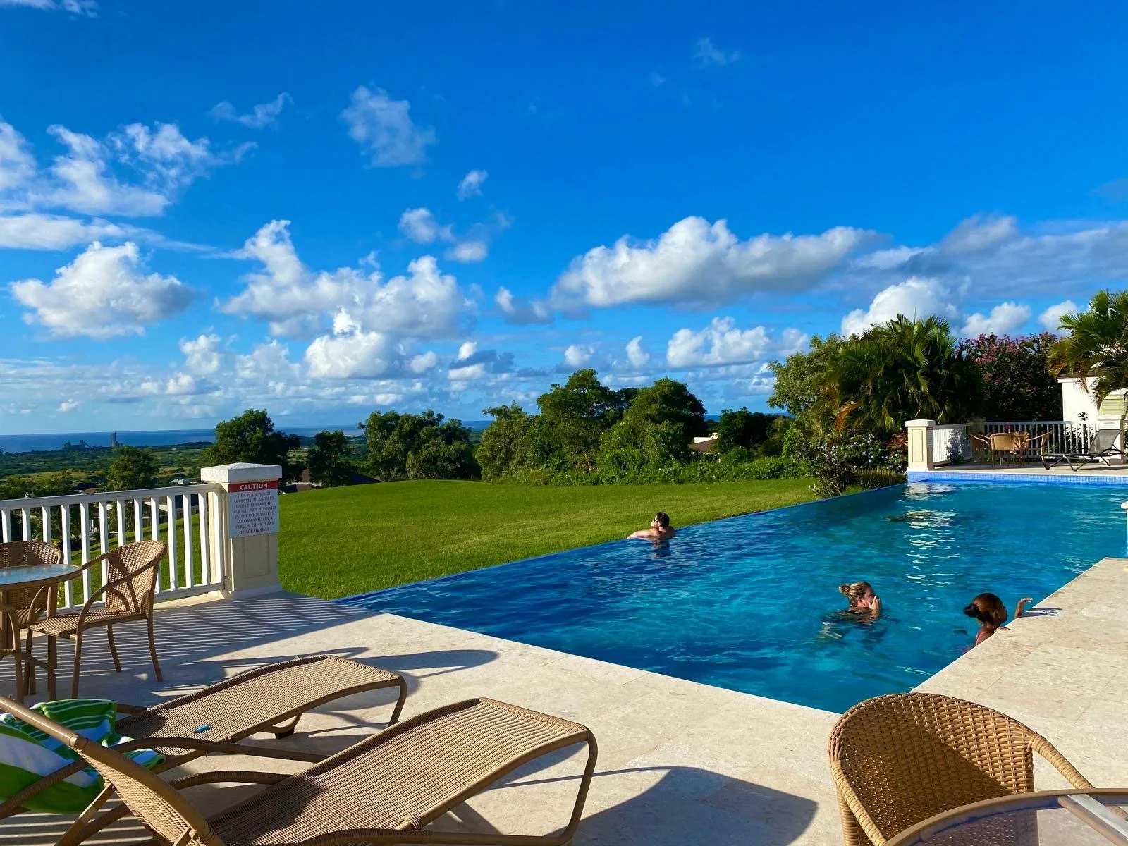 People swimming in an outdoor swimming pool with a scenic view of green trees, blue sky, and clouds in the background.