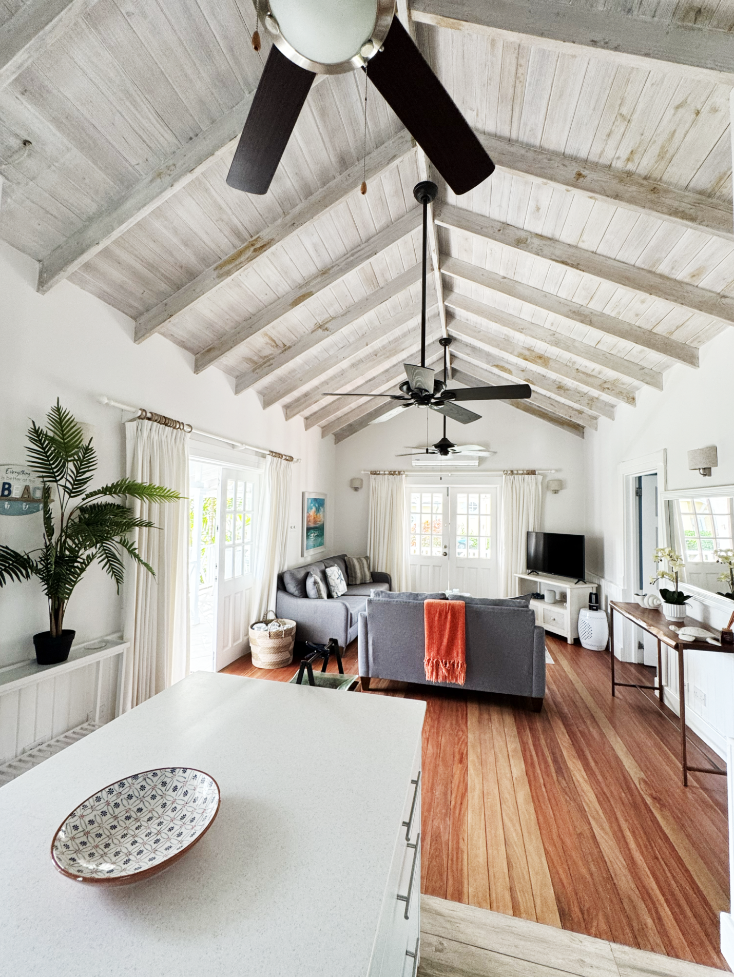 Living room with vaulted wooden ceiling, ceiling fans, beige curtains, grey sofas, a TV on a white console, indoor plants, and wooden flooring.