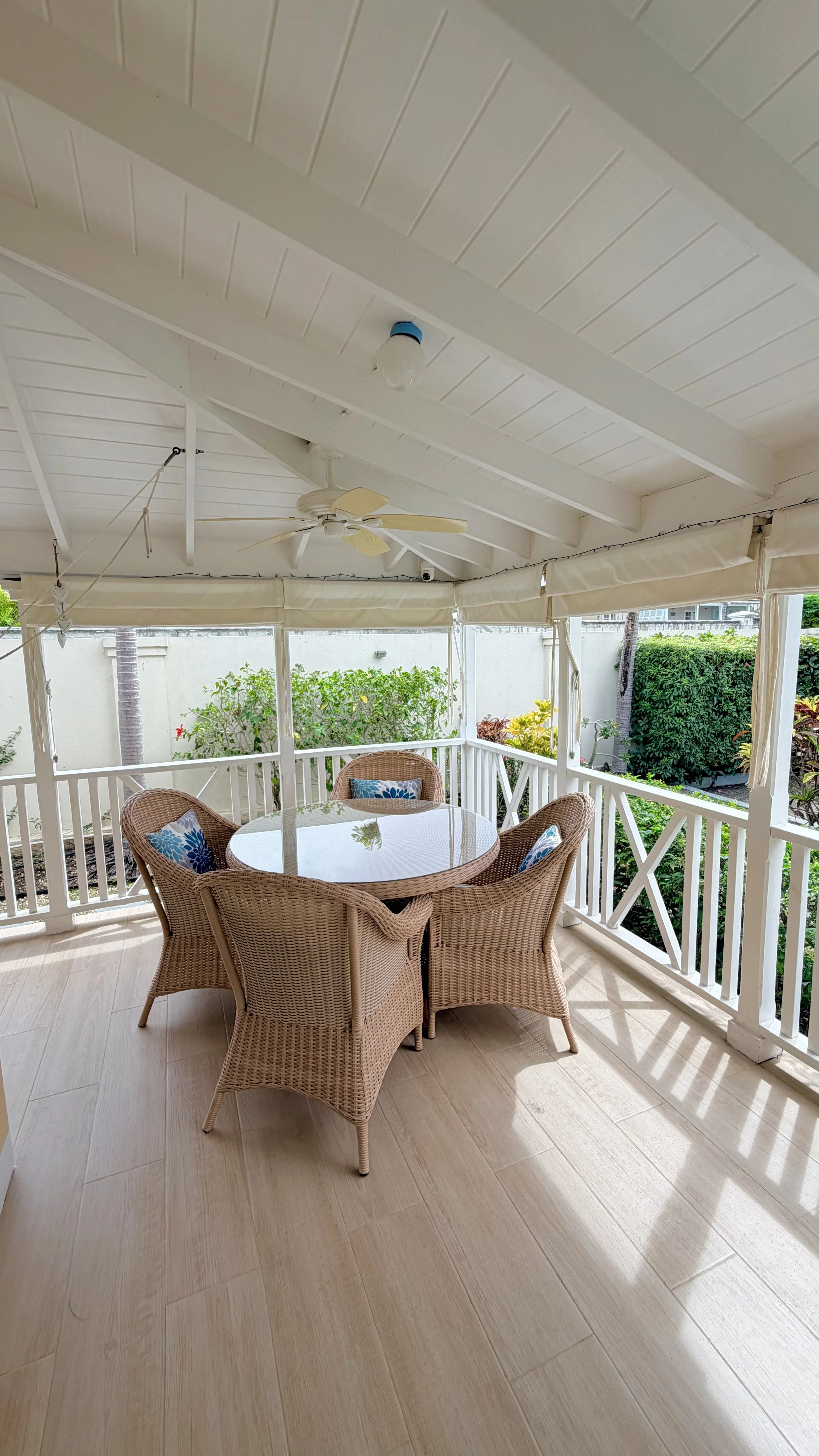 Covered outdoor patio area with a round glass table, four wicker chairs with blue patterned cushions, and a ceiling fan. The patio has a white railing and surrounding greenery.