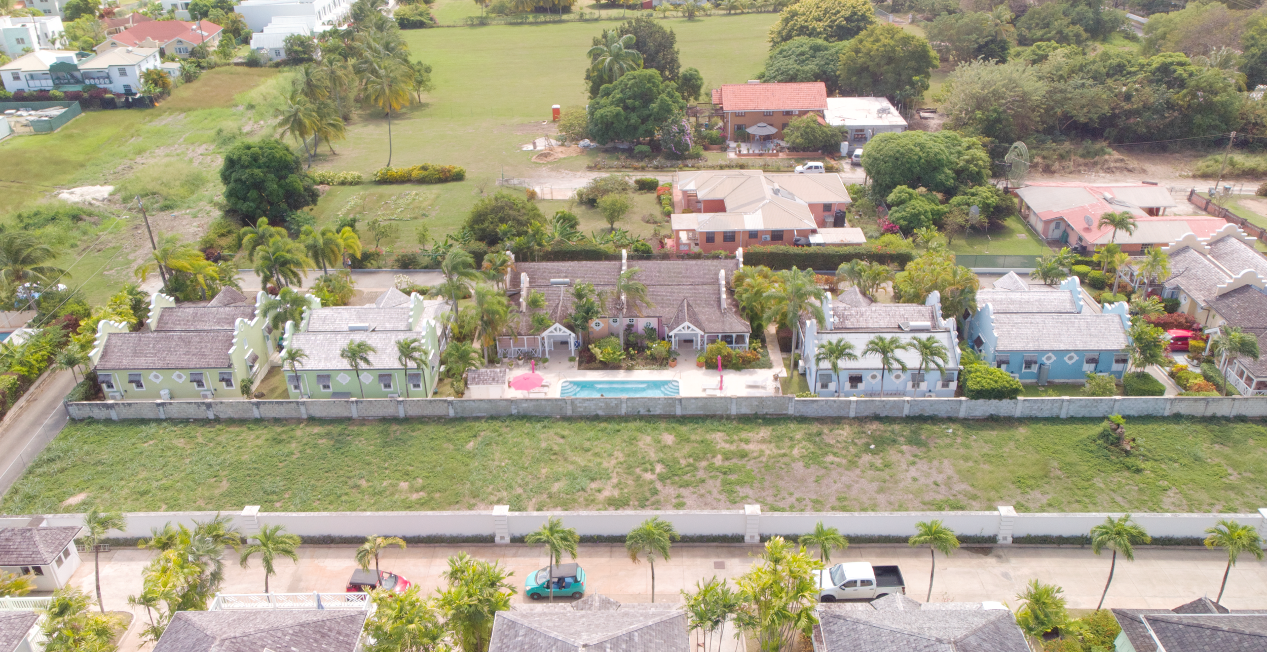Aerial view of a neighborhood with a row of colorful houses featuring a swimming pool, surrounded by lush greenery and trees.