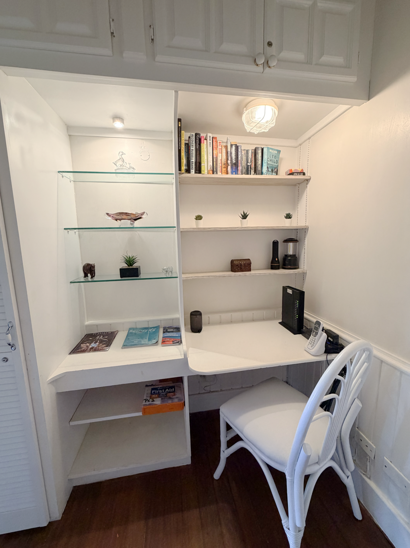 A small workspace with a white desk, two white chairs, and a built-in white bookshelf with three shelves holding plants, books, and decorative items. There is a telephone, cordless phone, and a wireless router on the desk. The overall decor is white 