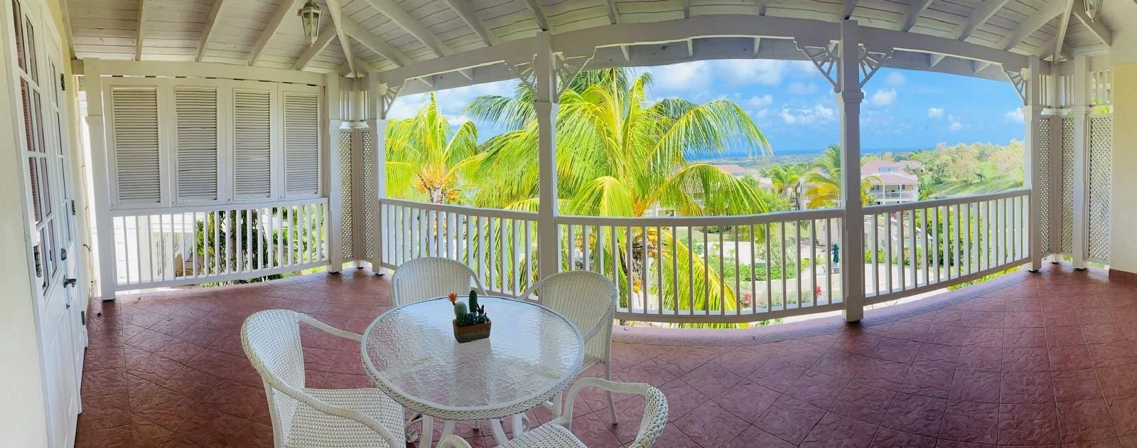 Panoramic view from a covered porch overlooking tropical palm trees, residential houses, and the ocean in the distance.