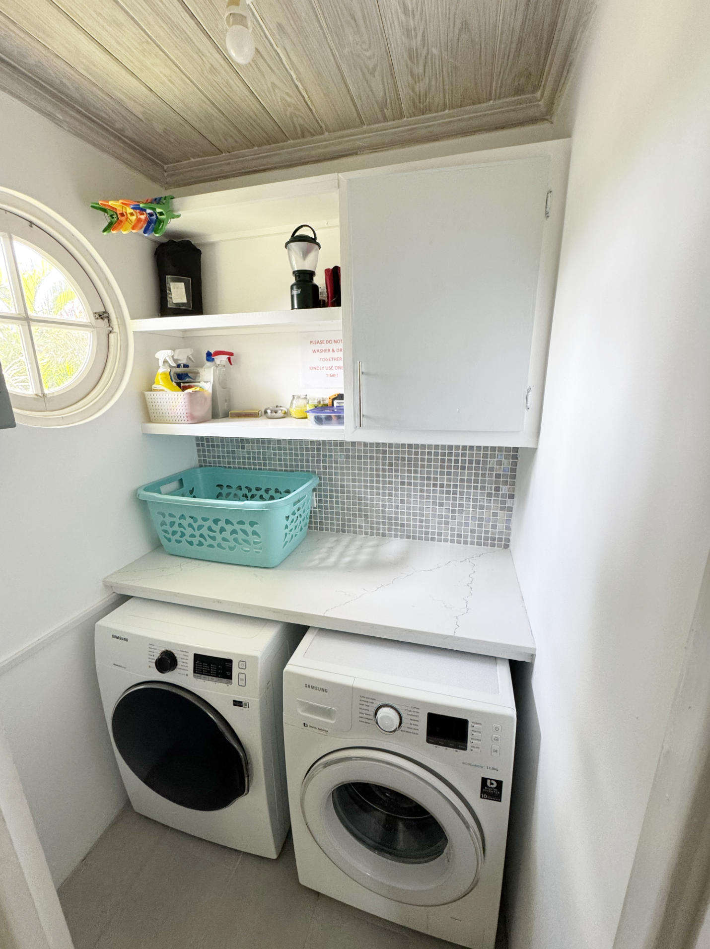 A laundry room with a washer and dryer, a white countertop with a teal laundry basket, white shelves with cleaning supplies, and a small window.