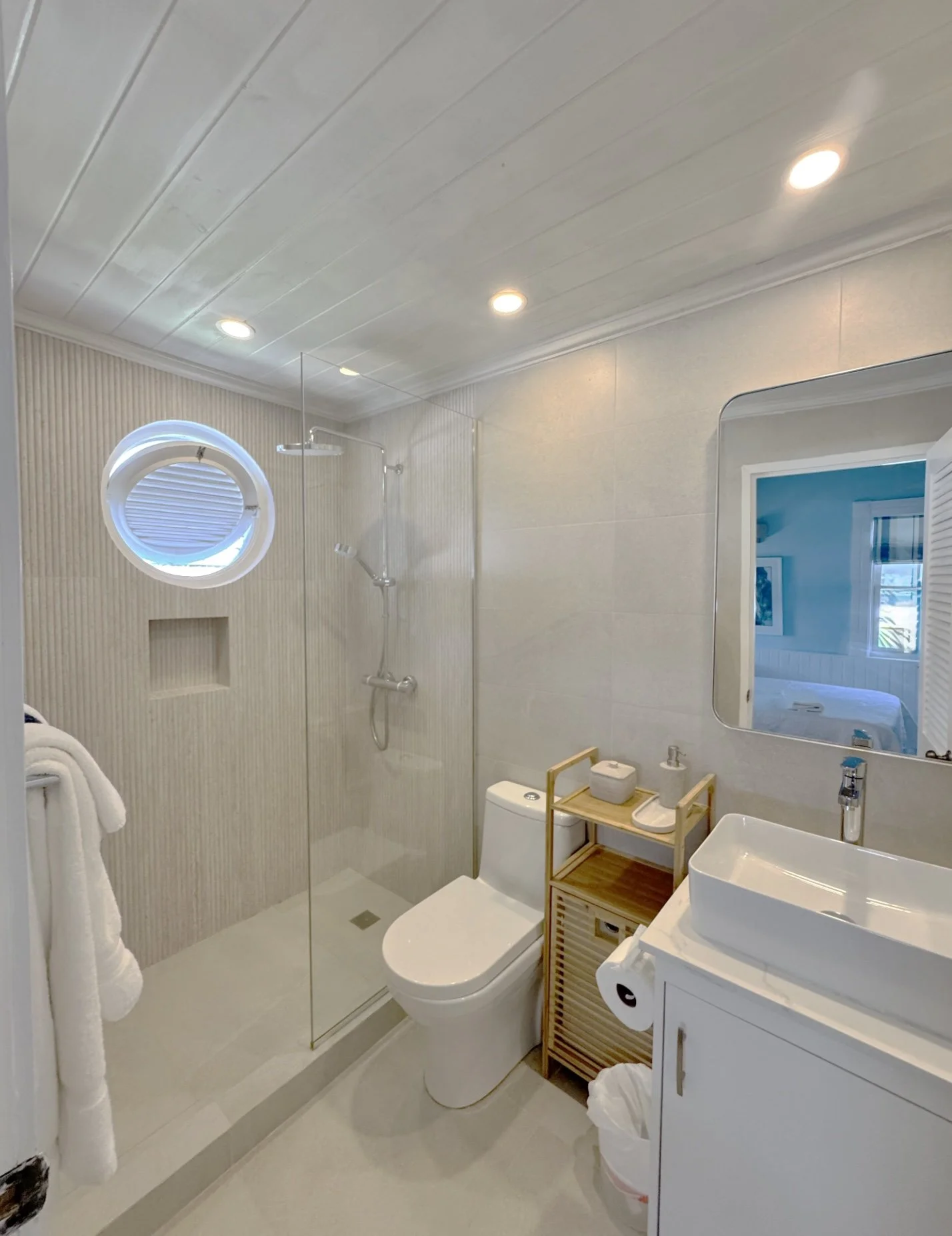 A modern bathroom with a glass shower enclosure, a white toilet, a wall-mounted sink with a white basin, a mirror, and a small wooden cabinet. The bathroom has light-colored walls, recessed ceiling lights, and a window with a blue roller shade.
