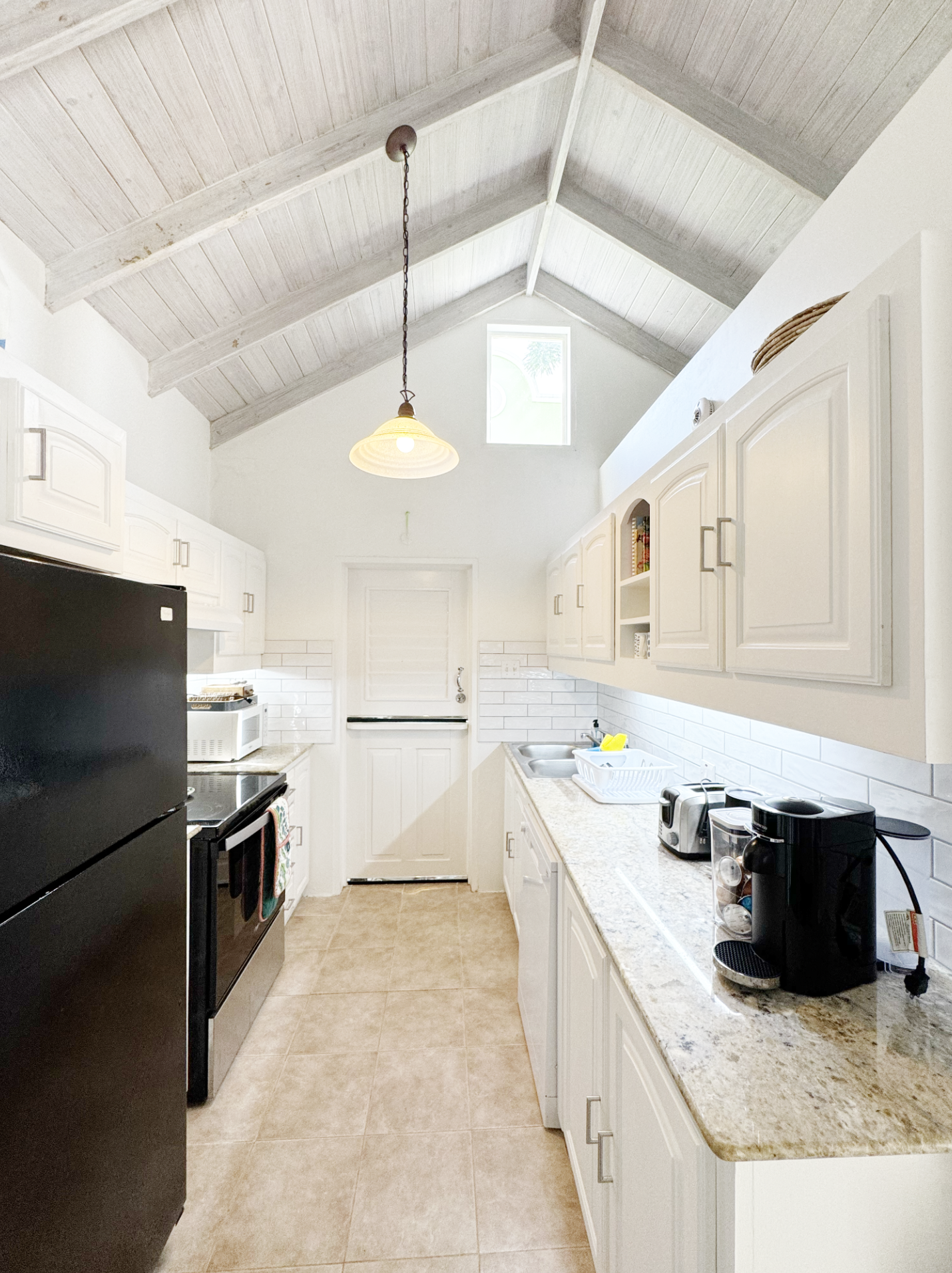 A small kitchen with white cabinets, beige granite countertop, black refrigerator, white stove, microwave, and coffee maker. The ceiling is vaulted with light wood paneling and a hanging light fixture. There is a small window and tile flooring.