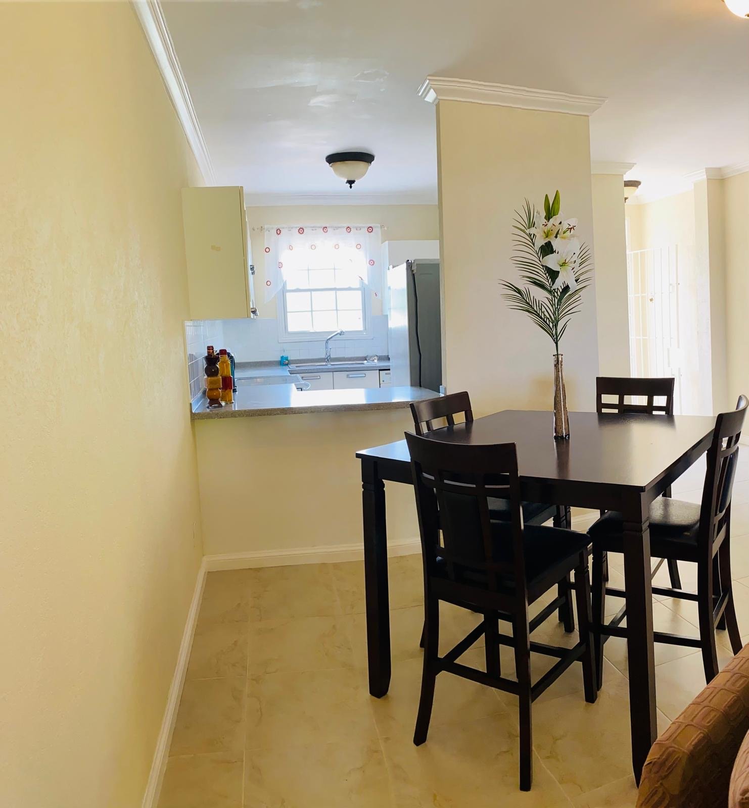 Dining area with black table and chairs, large flower vase with lilies, view of kitchen with window and countertop.