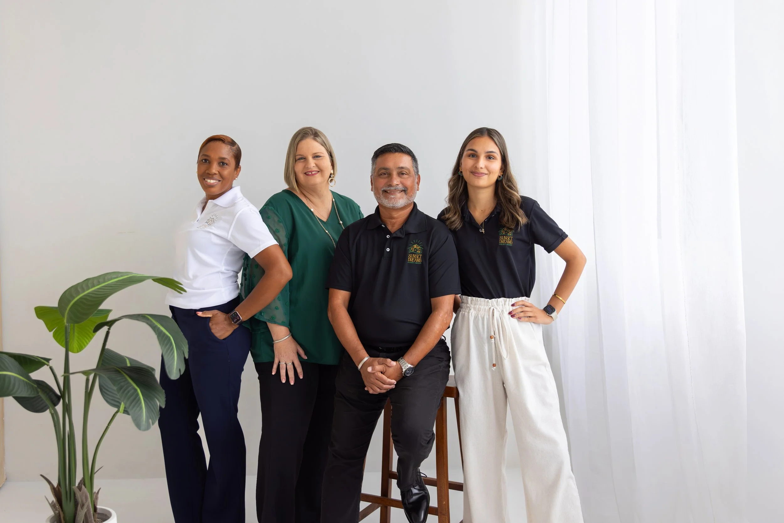 Four diverse individuals standing close together in front of a plain white wall, smiling and posing for the camera, with a potted plant visible in the foreground.
