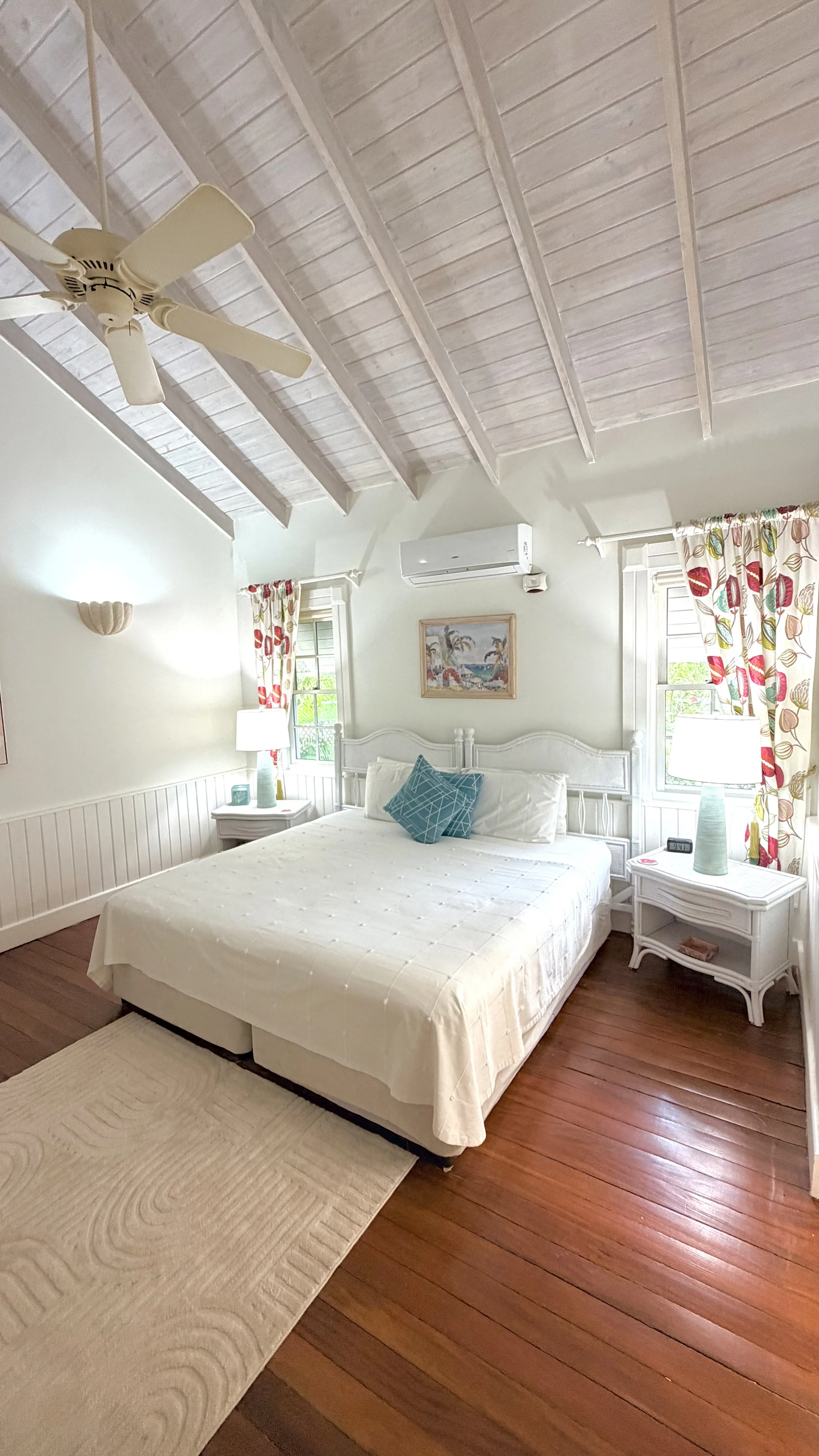 A bright bedroom with a white bed, side tables, lamps, a wall-mounted air conditioner, and windows with floral curtains. The ceiling is vaulted with white wooden beams and a ceiling fan.