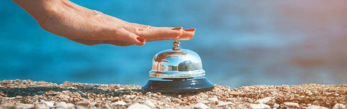 A person pressing a service bell on a pebble surface near water.