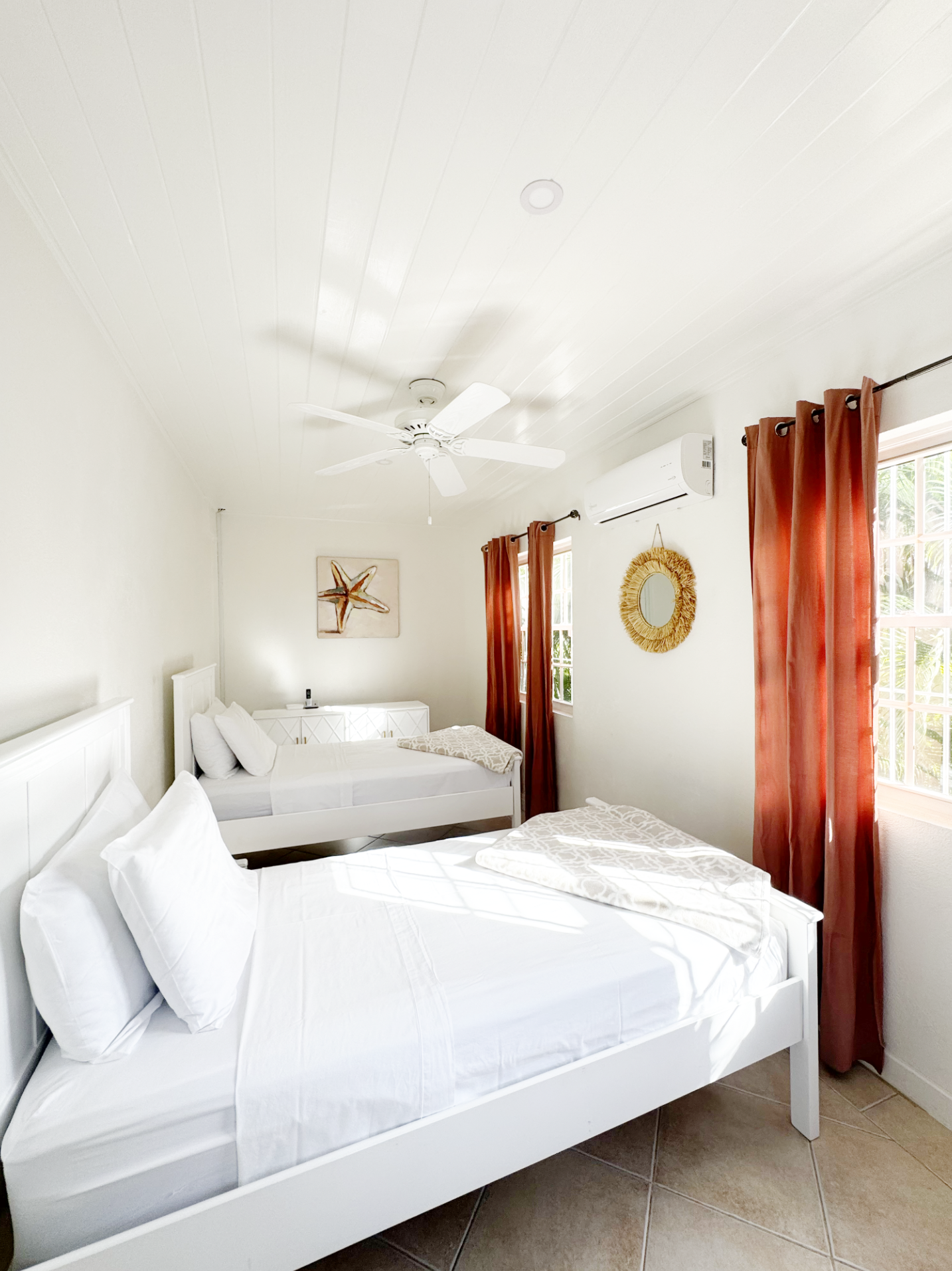 A bright bedroom with white walls and ceiling, featuring two white beds with pillows and folded blankets. There is a small white nightstand beside the beds, a starfish artwork on the wall, and a round mirror with a straw frame. The room has two windo