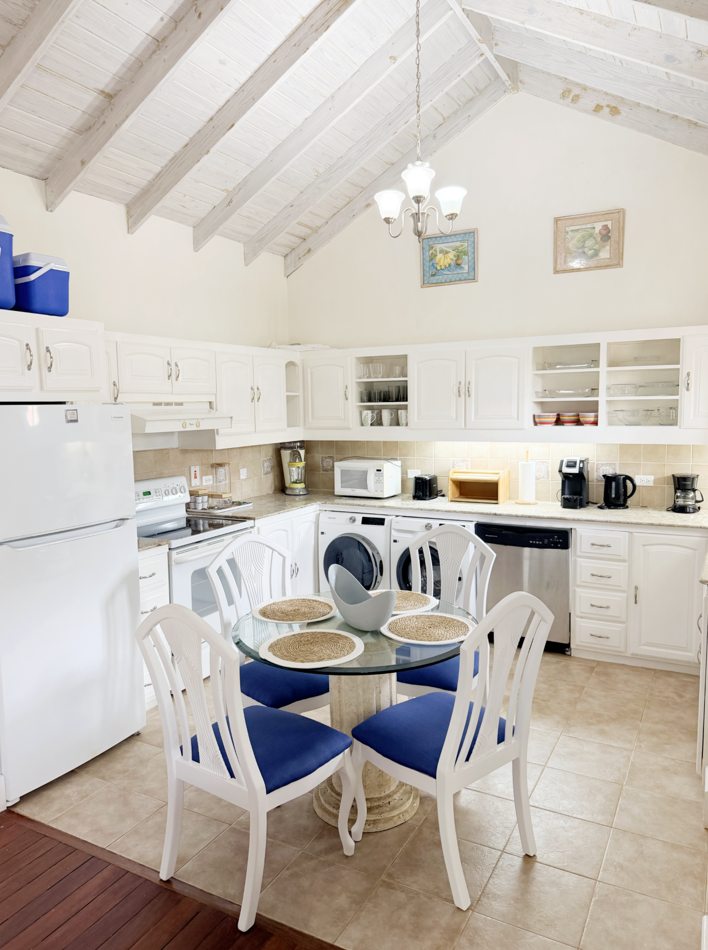 Kitchen with white cabinets, a round dining table with four chairs, and various appliances including a refrigerator, microwave, and coffee maker. The ceiling is vaulted with exposed wood beams and a chandelier hanging from it.