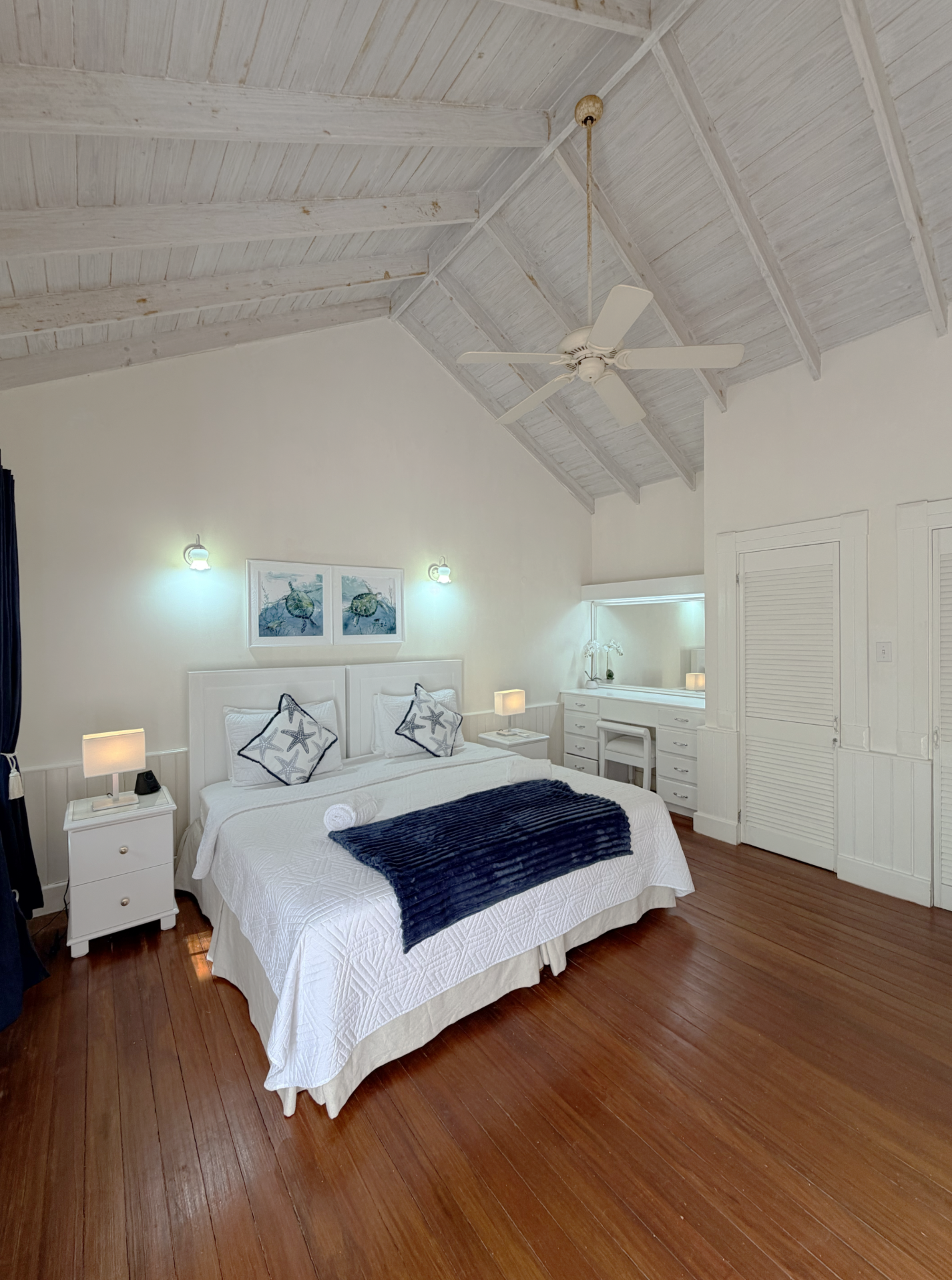 A bedroom with a white bed, navy blanket, and starfish pillows. The room has wooden flooring, white walls, and a high, painted, vaulted ceiling with a ceiling fan. There are two small nightstands with lamps and a framed sea-themed artwork above the b