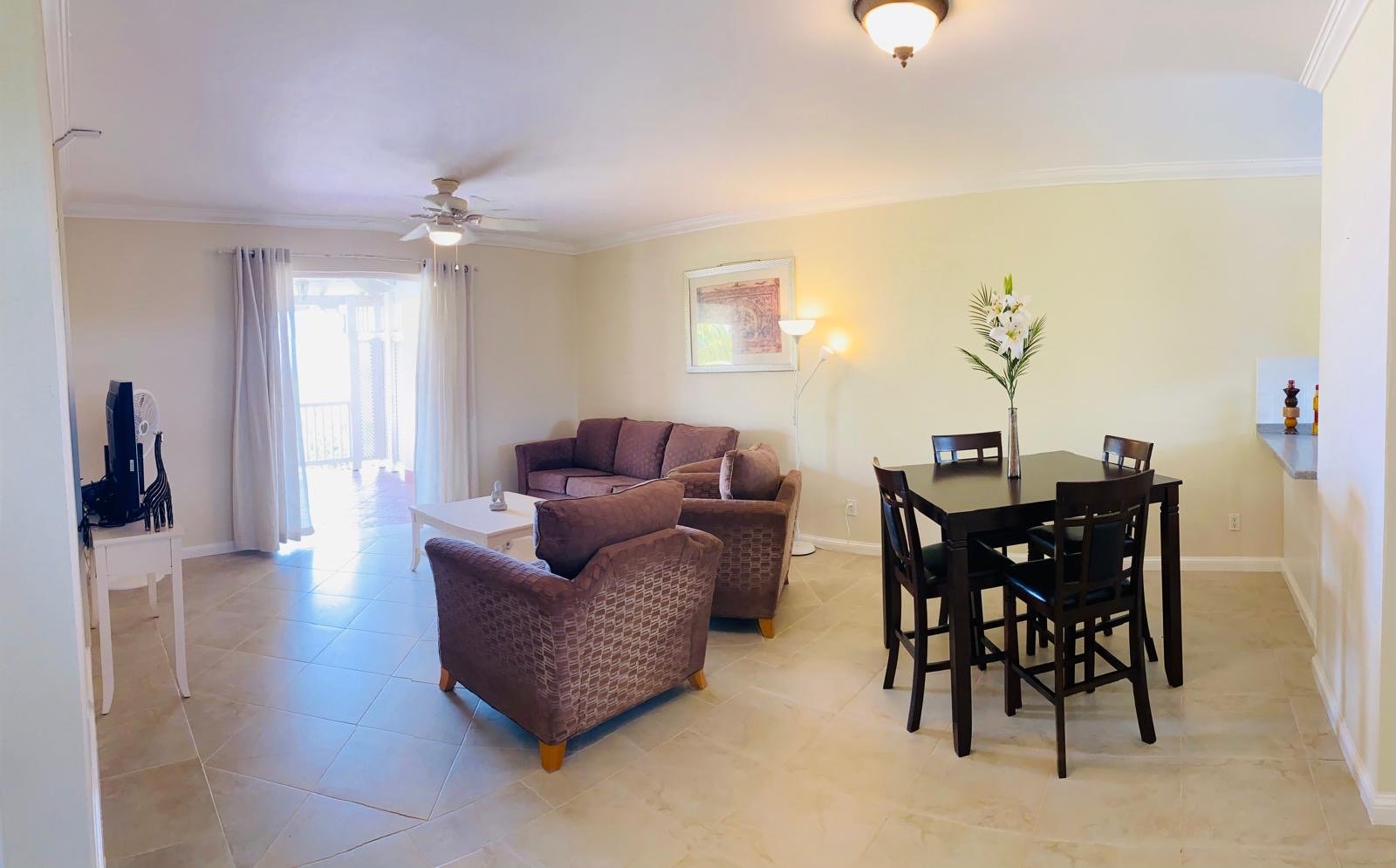 Living room with beige tiled floor, beige walls, a ceiling fan with light, a brown sofa, two brown armchairs, a white coffee table, a black dining table with four chairs, a tall white standing lamp, a framed picture on the wall, a vase with white flo