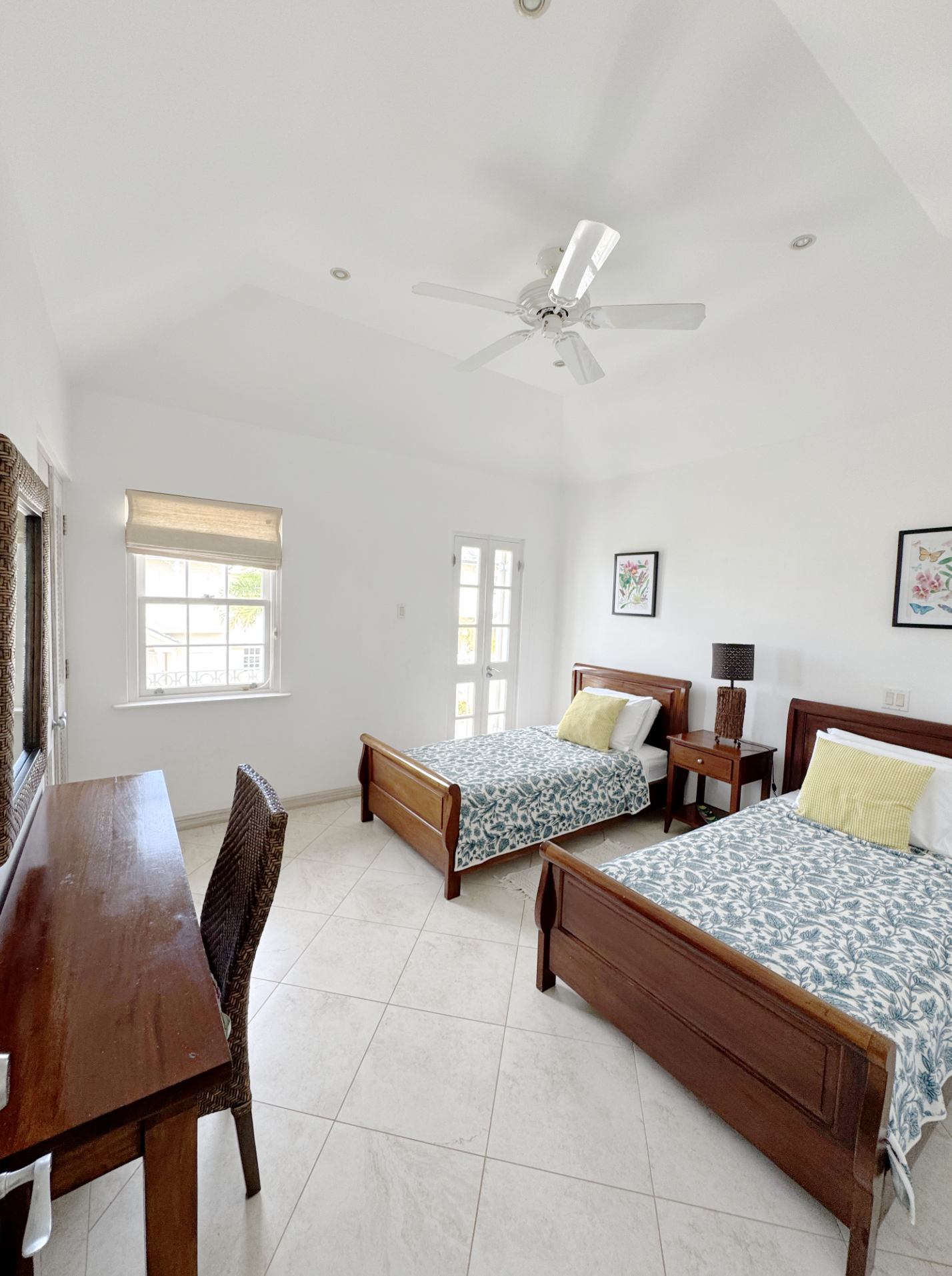 A bright bedroom with two wooden beds featuring blue and white patterned bedding and yellow pillows, two framed floral pictures on the wall, a window with a beige shade, a wooden nightstand with a black lamp, a ceiling fan, and a wooden dresser with 