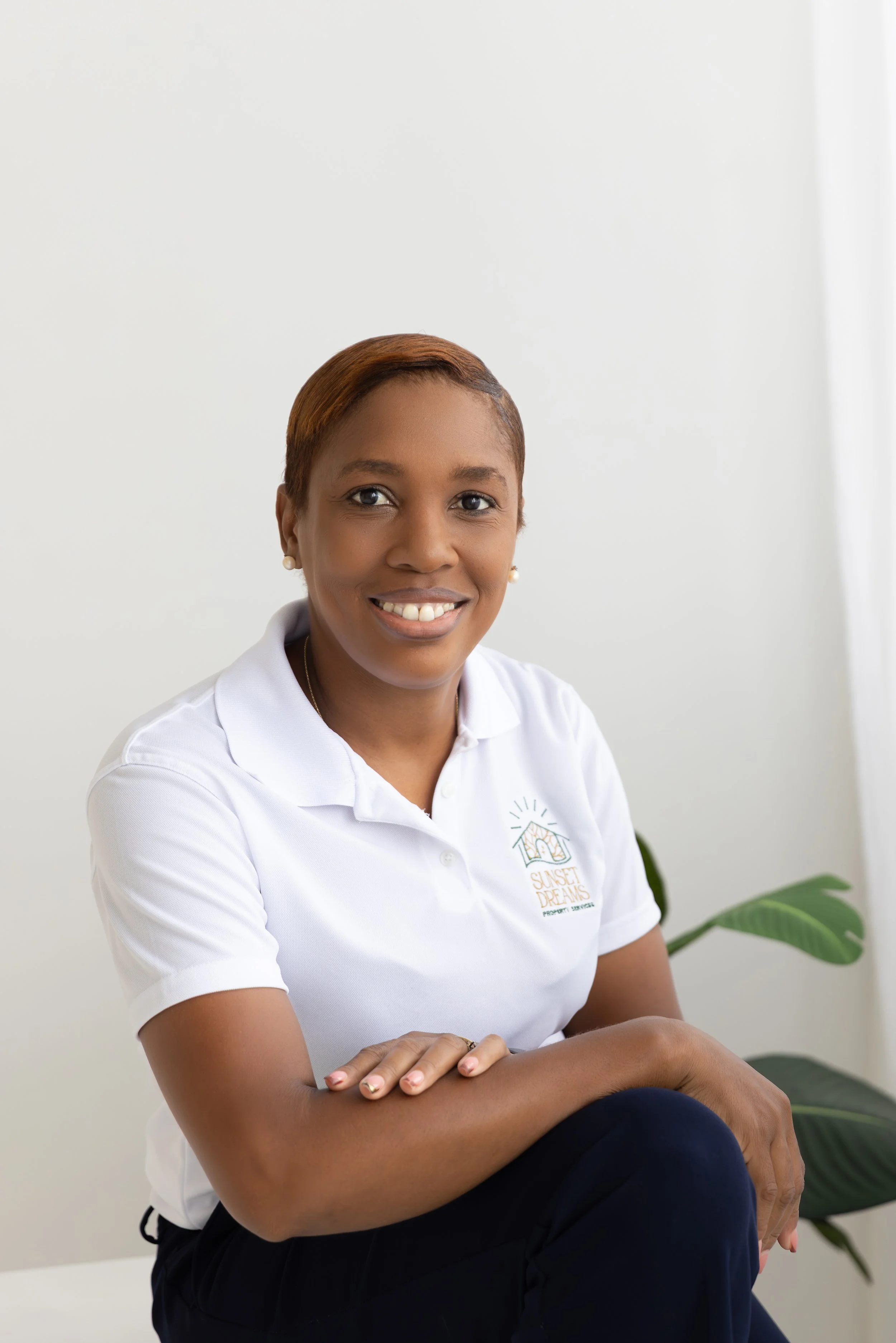 A young woman with short, reddish-brown hair sitting cross-legged with her arms resting on her knees, smiling at the camera, wearing a white polo shirt with a logo that reads 'Sunset Dreams Property Management,' in a room with white walls and a large green plant beside her.